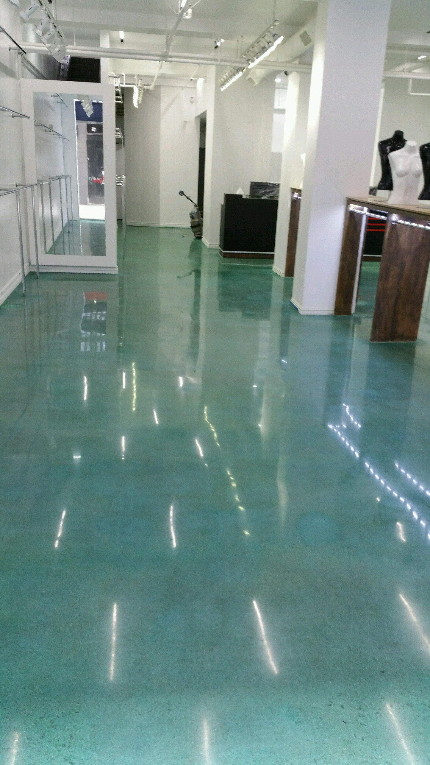 Empty retail store with green polished floor, white walls, mannequins on displays, and metal clothing racks.
