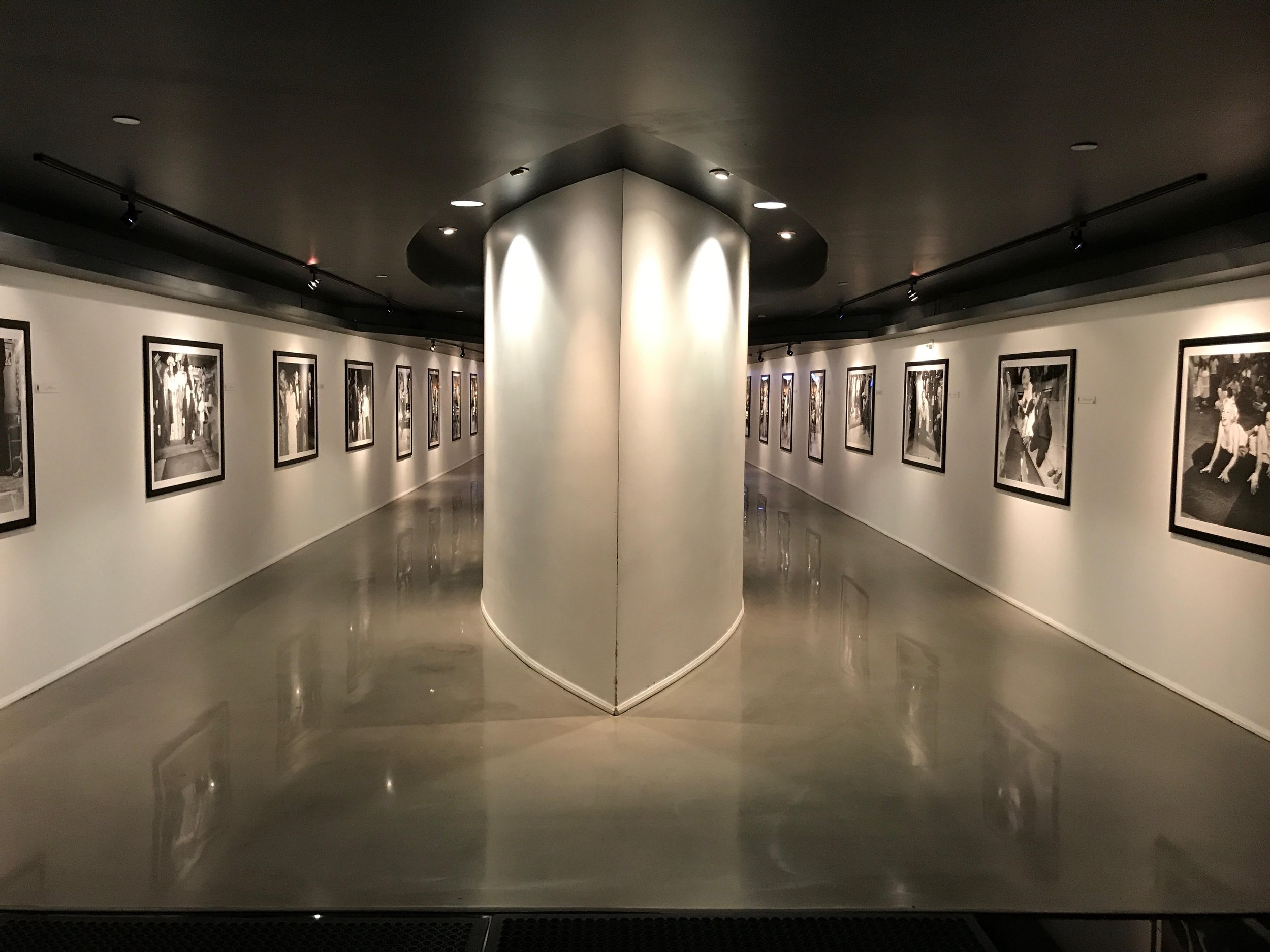 An art gallery with framed pictures displayed on white walls, a central white pillar, and a reflective gray floor.