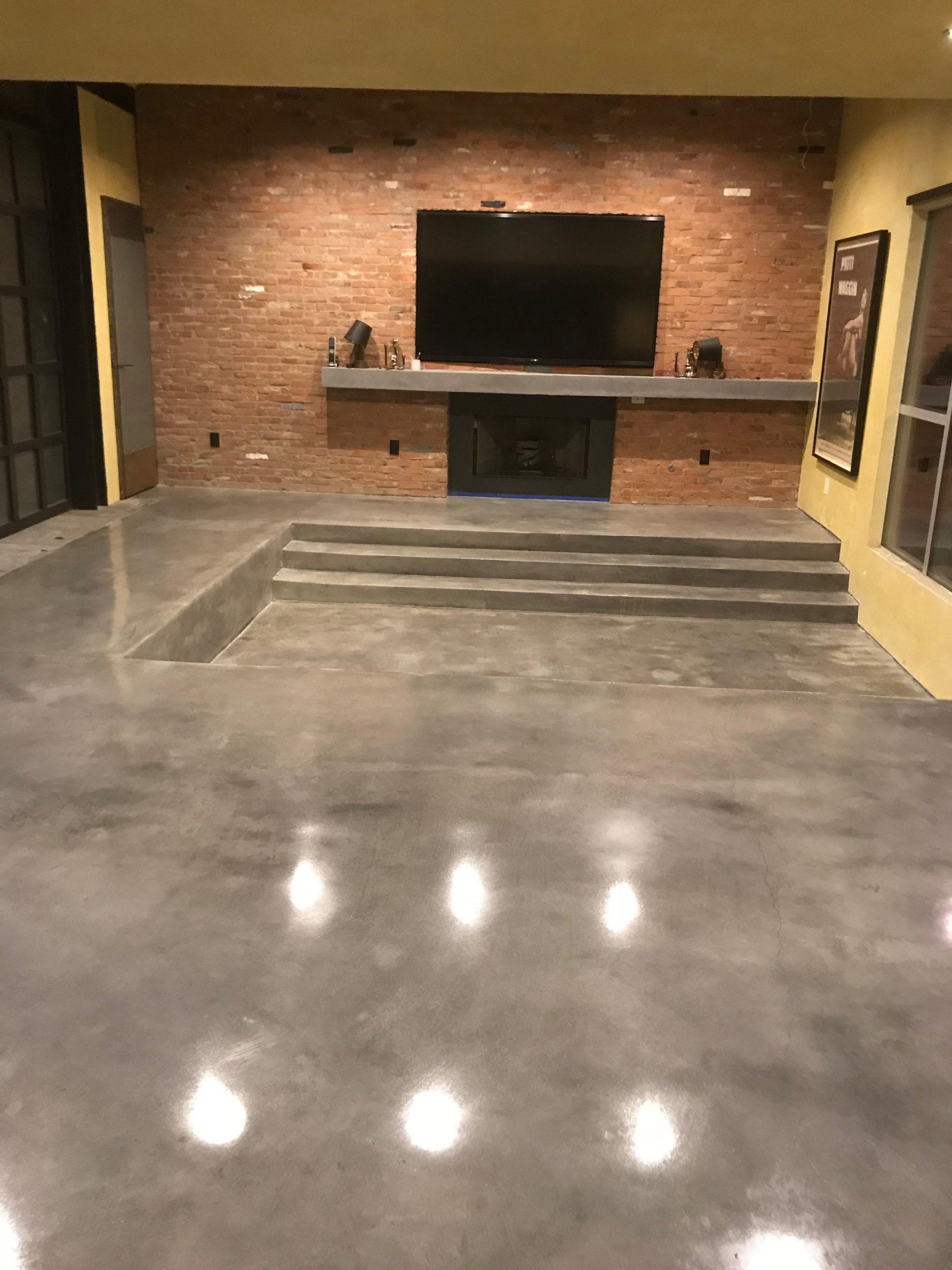 Modern living room with polished concrete floor, brick wall, mounted flat-screen TV, and a fireplace with surrounding benches.