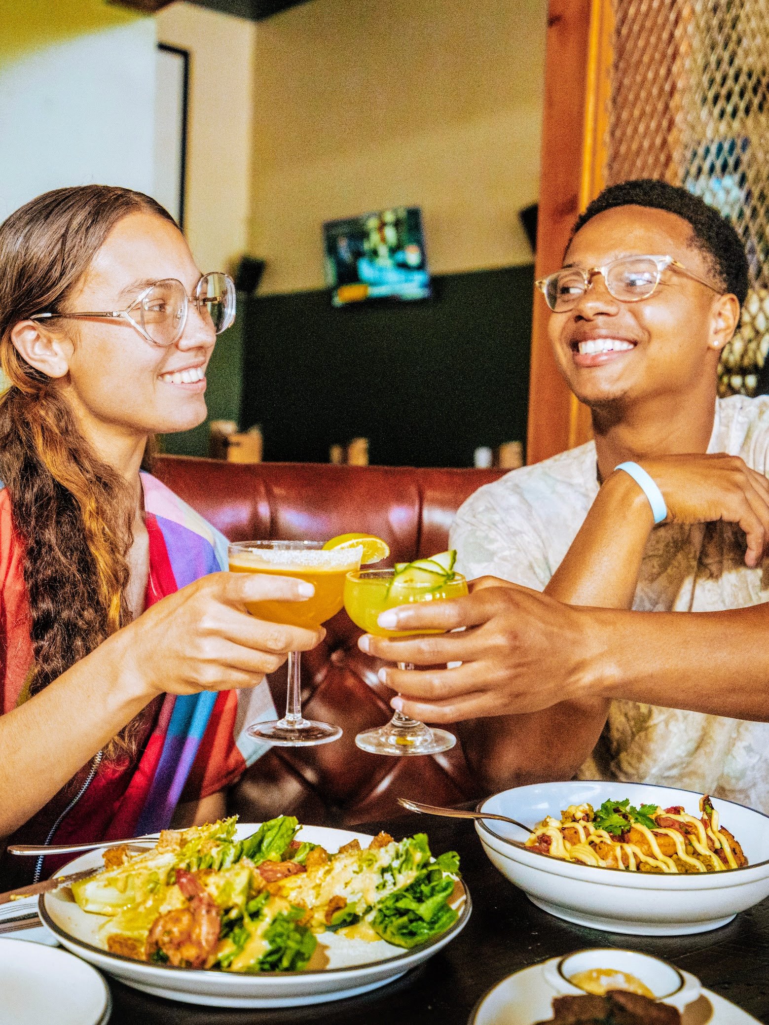 Two women clinking cocktail glasses with lemon wedges at a dinner table in a restaurant, with plates of food including salad and pasta in front of them.