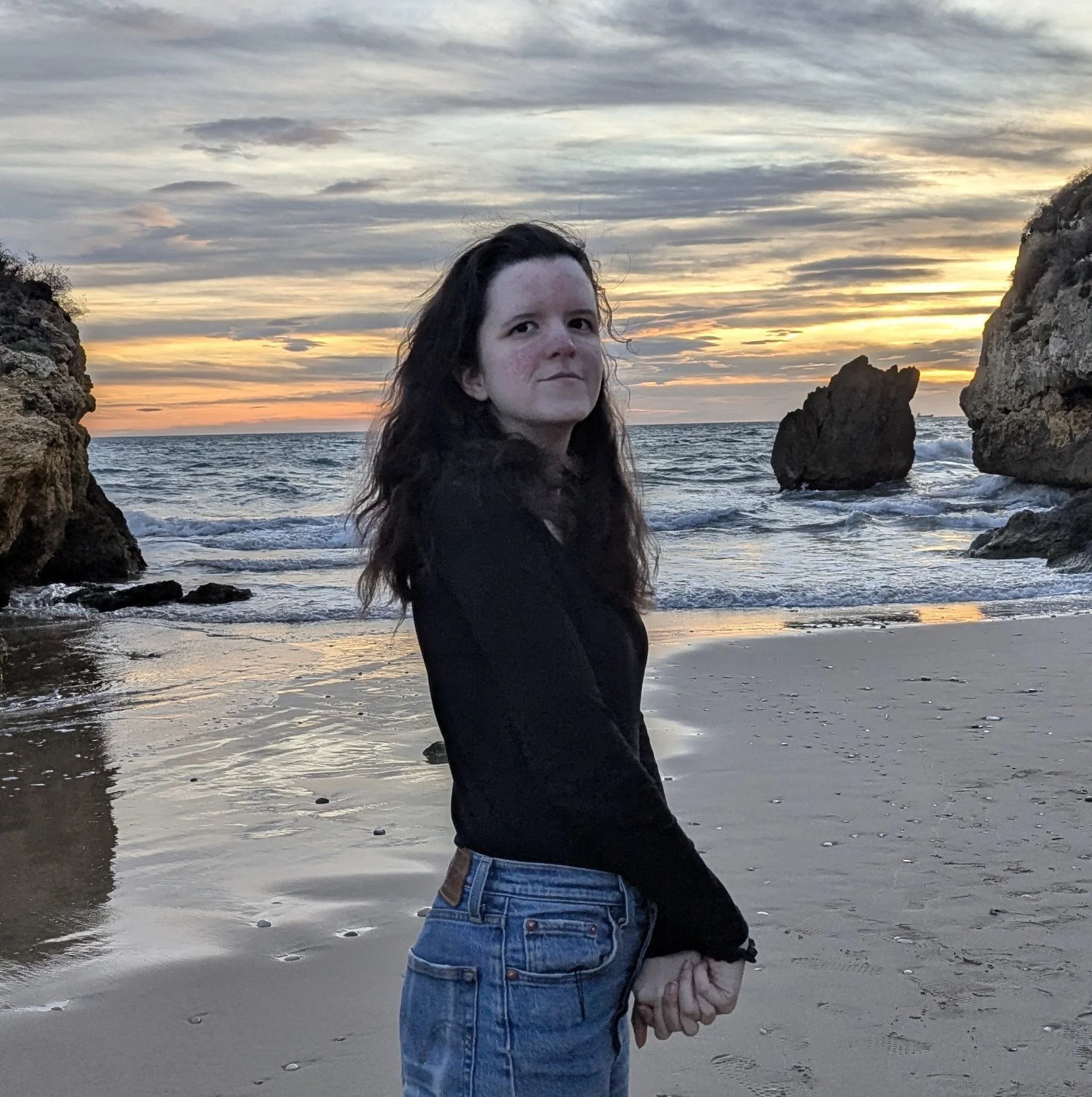 A woman with long dark hair stands on a beach during sunset, holding a seashell in her hand with large rocks and clouds in the sky behind her.
