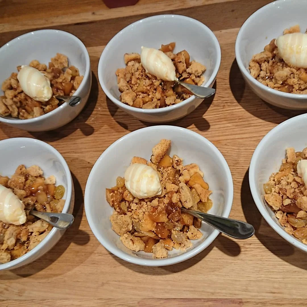 Six bowls of apple crumble dessert with cream on top, on a wooden table.