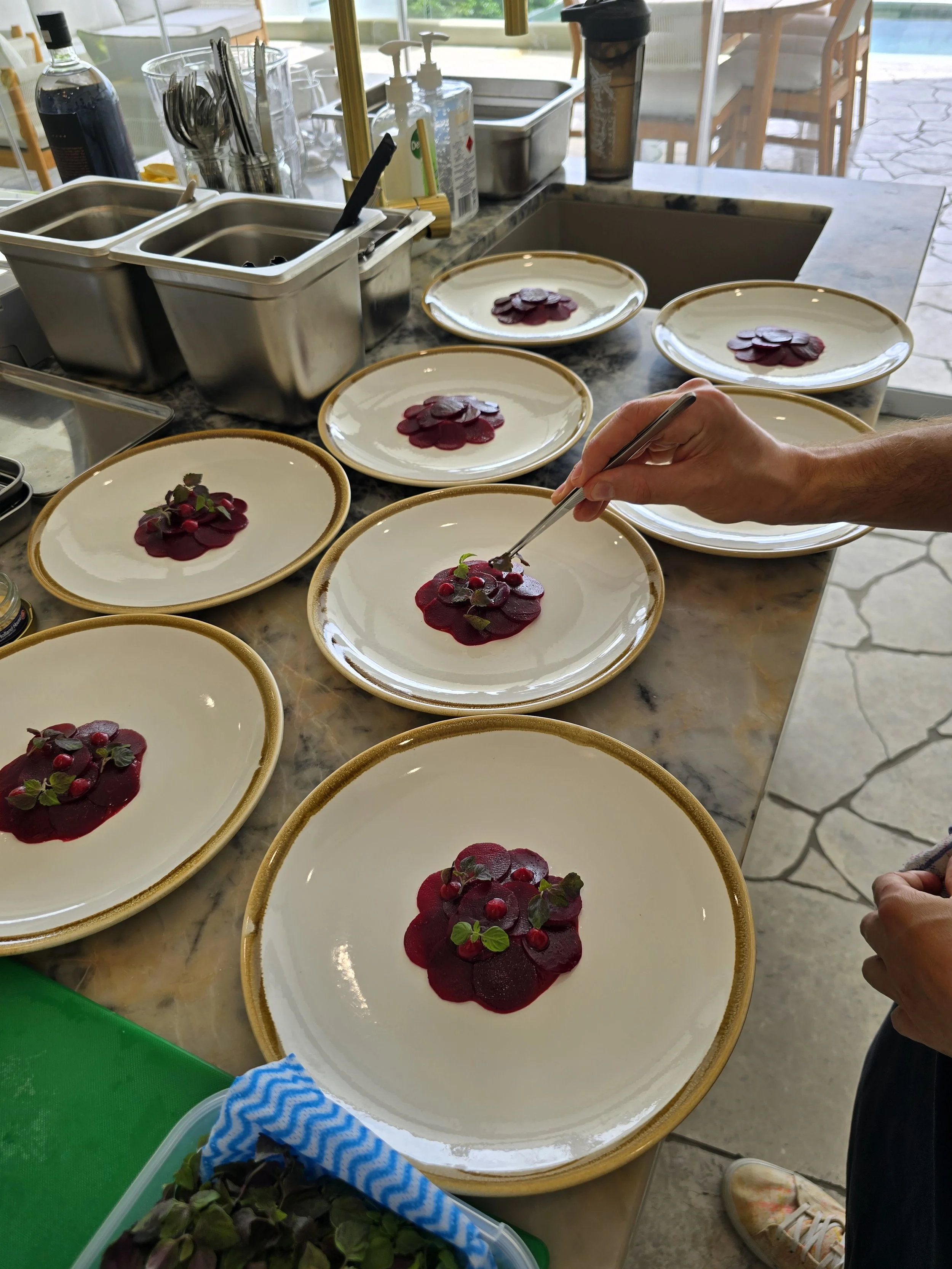 Chef plating beetroot carpaccio with microgreens on multiple plates in a kitchen setting.