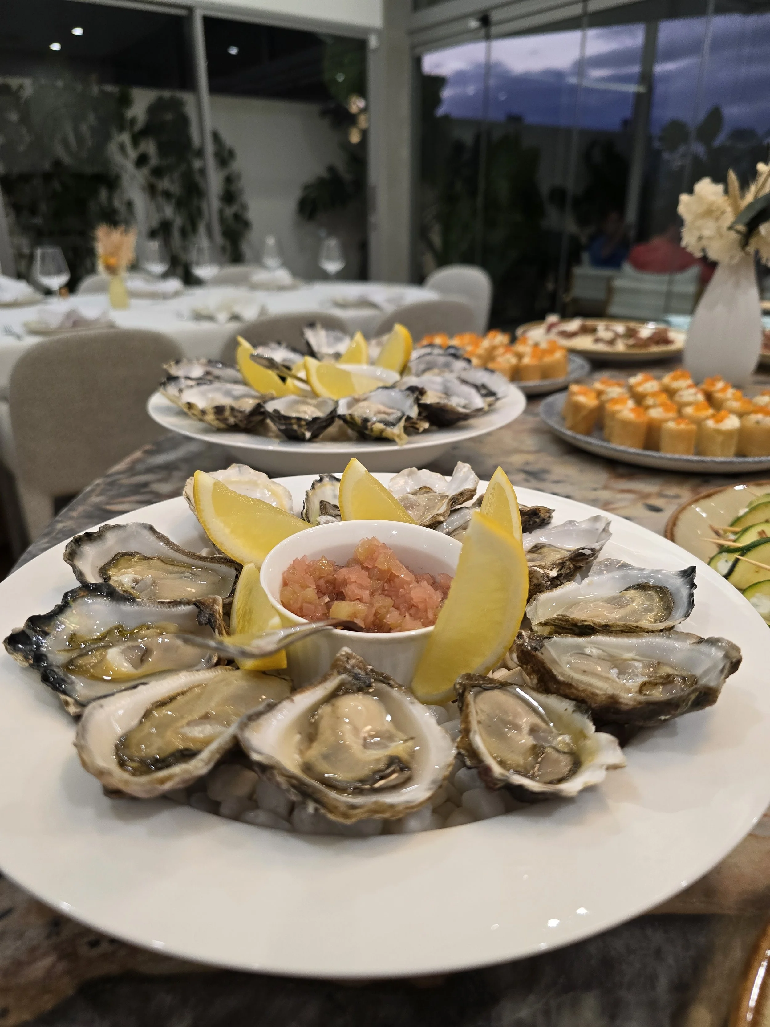 Plates of fresh oysters on a table with lemon wedges and mignonette sauce, surrounded by other appetizers in a dining setting.