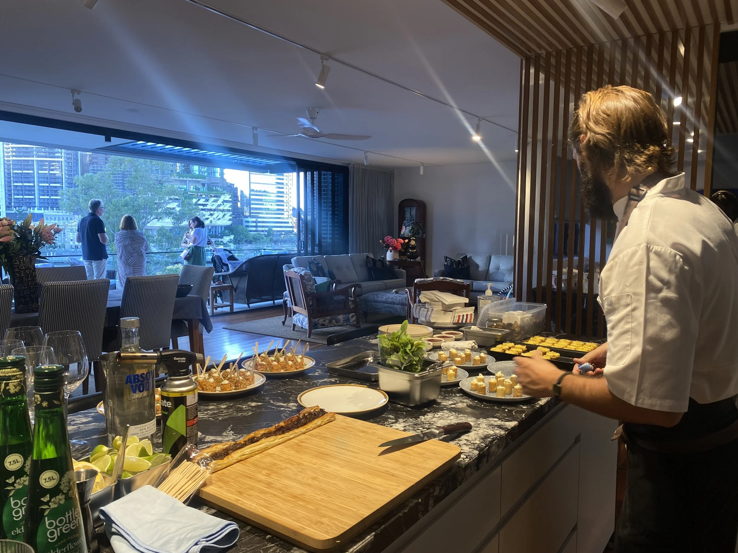 Chef preparing food in a modern kitchen with a view of an outdoor patio and cityscape. Several people are standing on the patio. The counter has ingredients, drinks, and appetizers.
