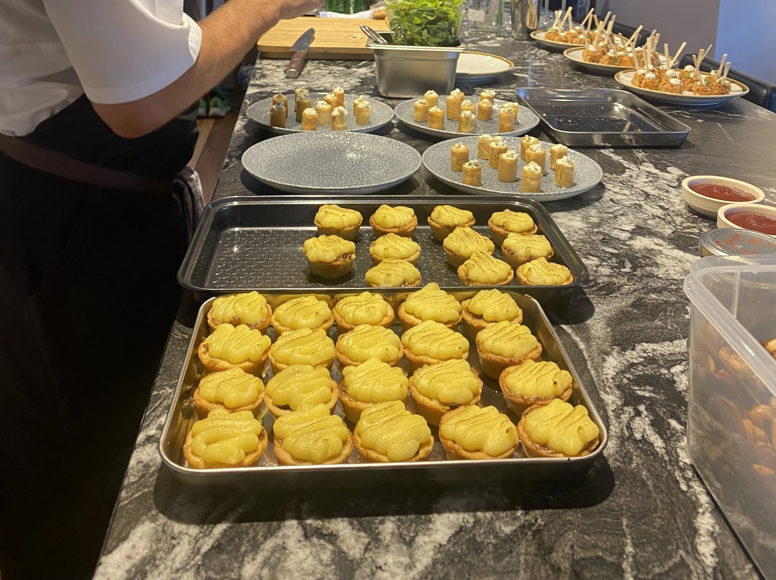 Chef preparing various appetizers in a kitchen, including pastry shells with mashed potatoes, stuffed rolls, and skewered meatballs on a marble countertop.