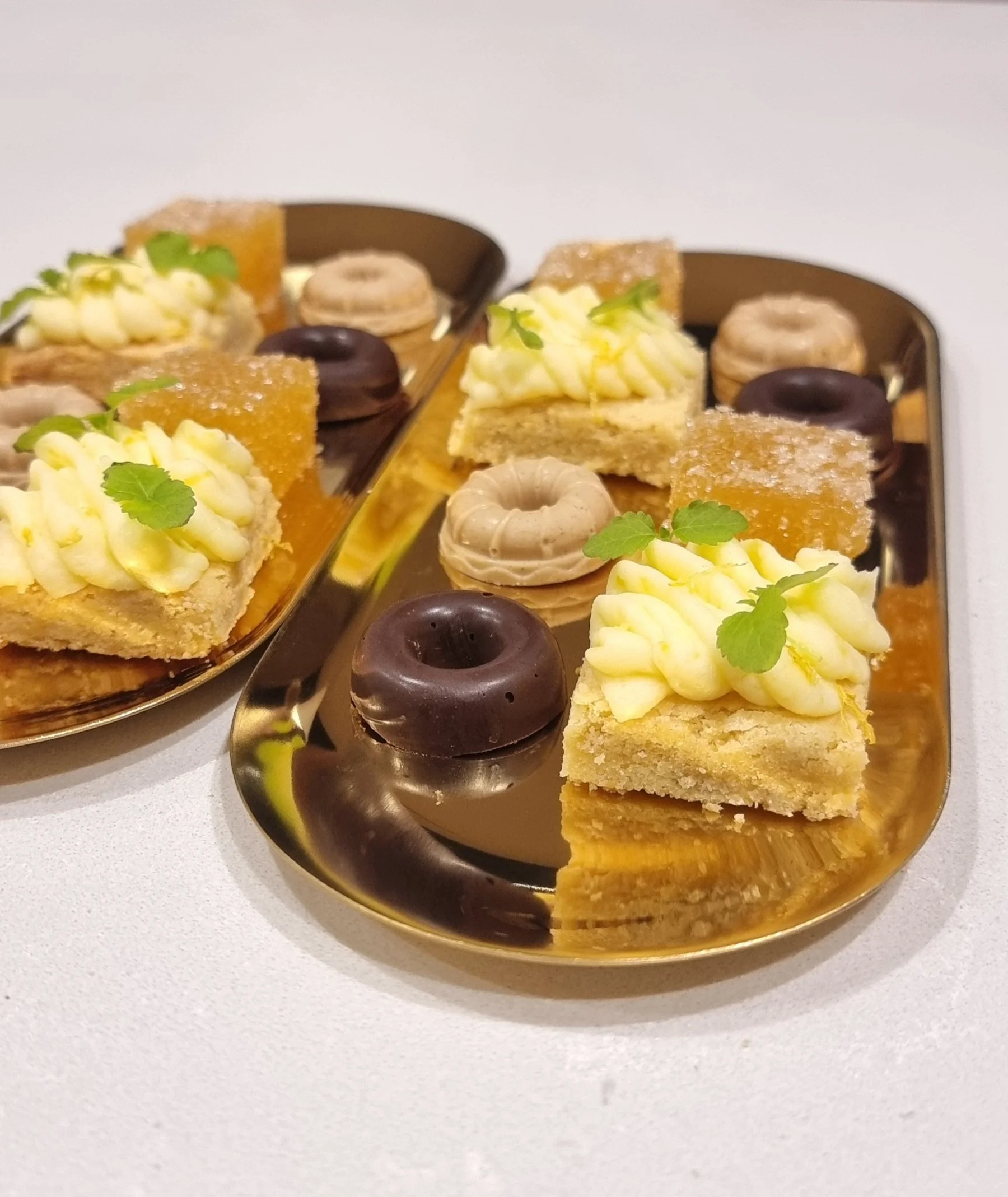 Dessert platter with assorted pastries, including squares with cream topping, small donuts, and jelly cubes, garnished with green herbs on gold trays.