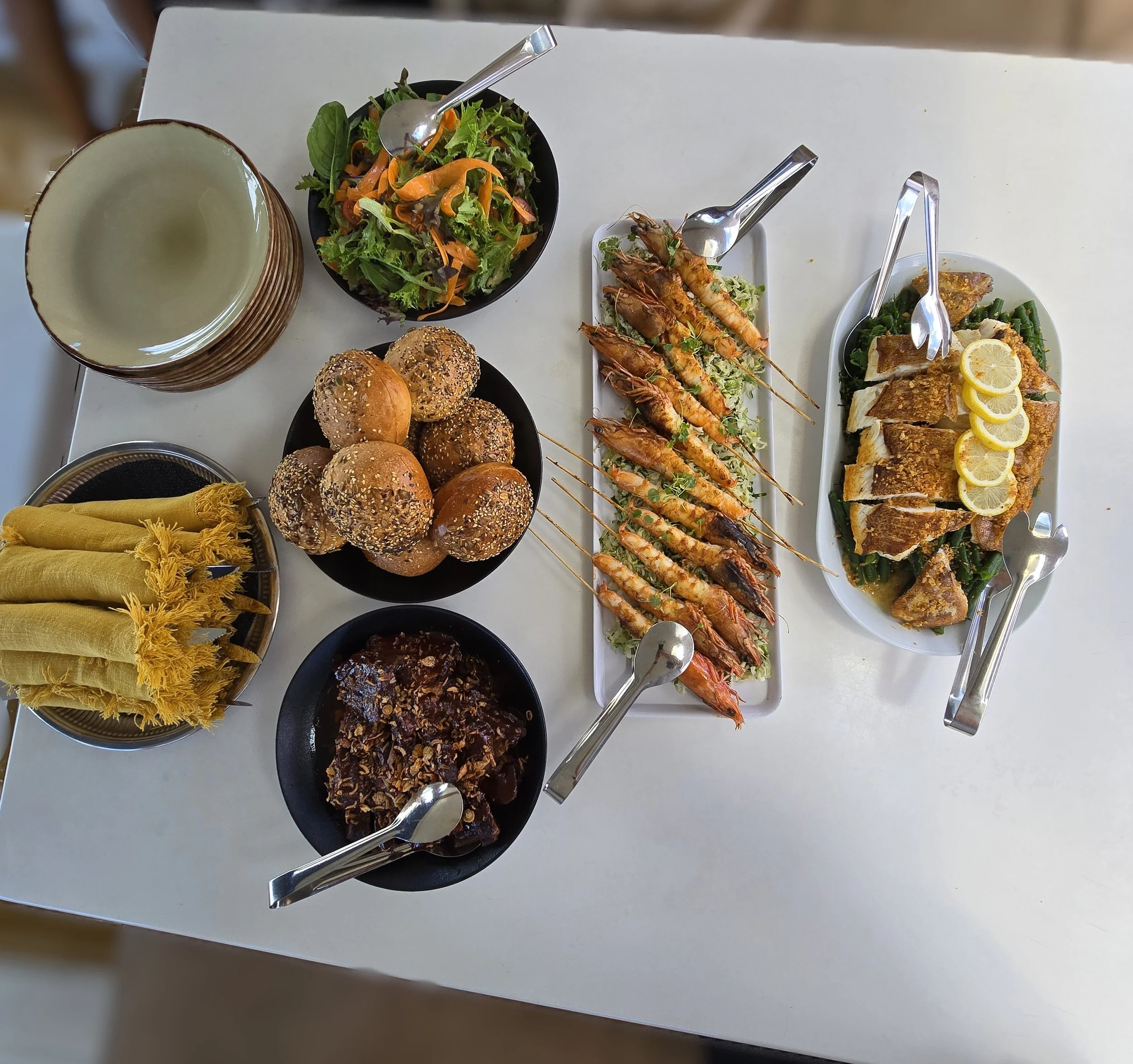 Table with plates of rolls, salad, skewered prawns, grilled fish with lemon, wrapped utensils, and a dish of cooked beef brisket.