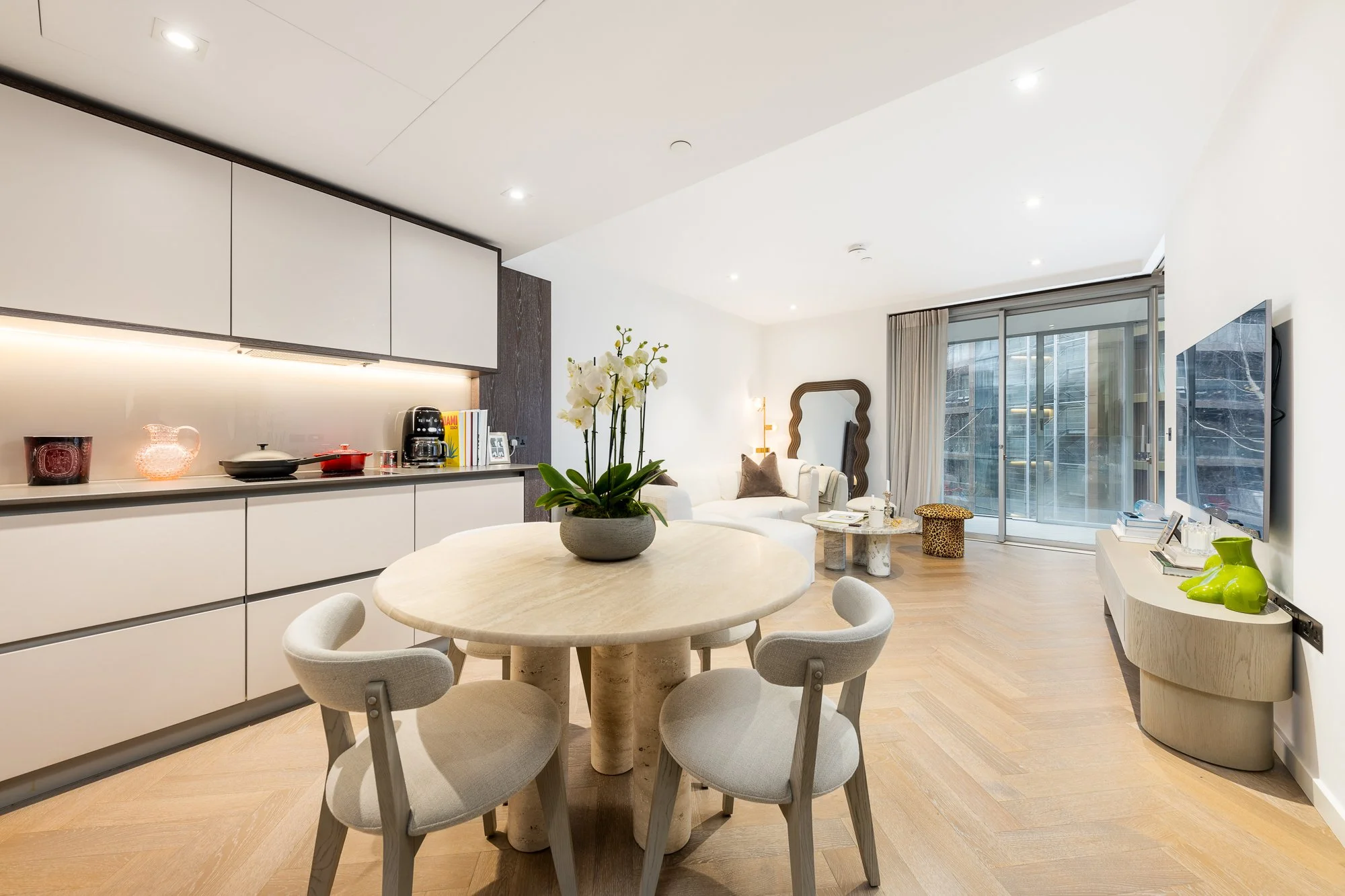 Modern living room and kitchen with dining table, white couch, TV, and large windows.