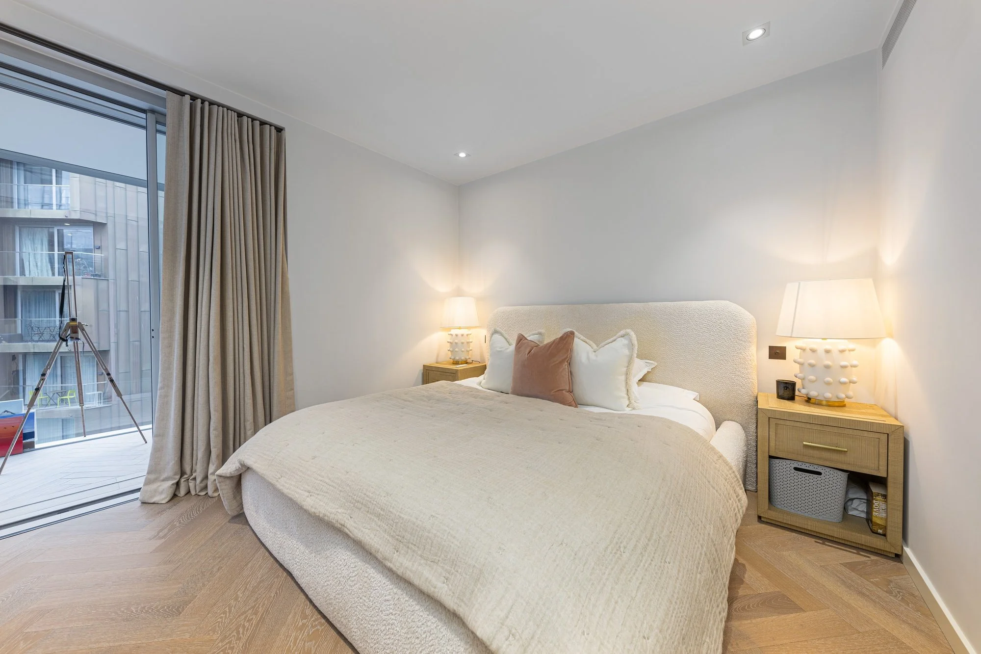 Modern bedroom with a large bed, beige bedding, decorative pillows, two bedside tables with lamps, and a sliding glass door with beige curtains leading to a balcony.