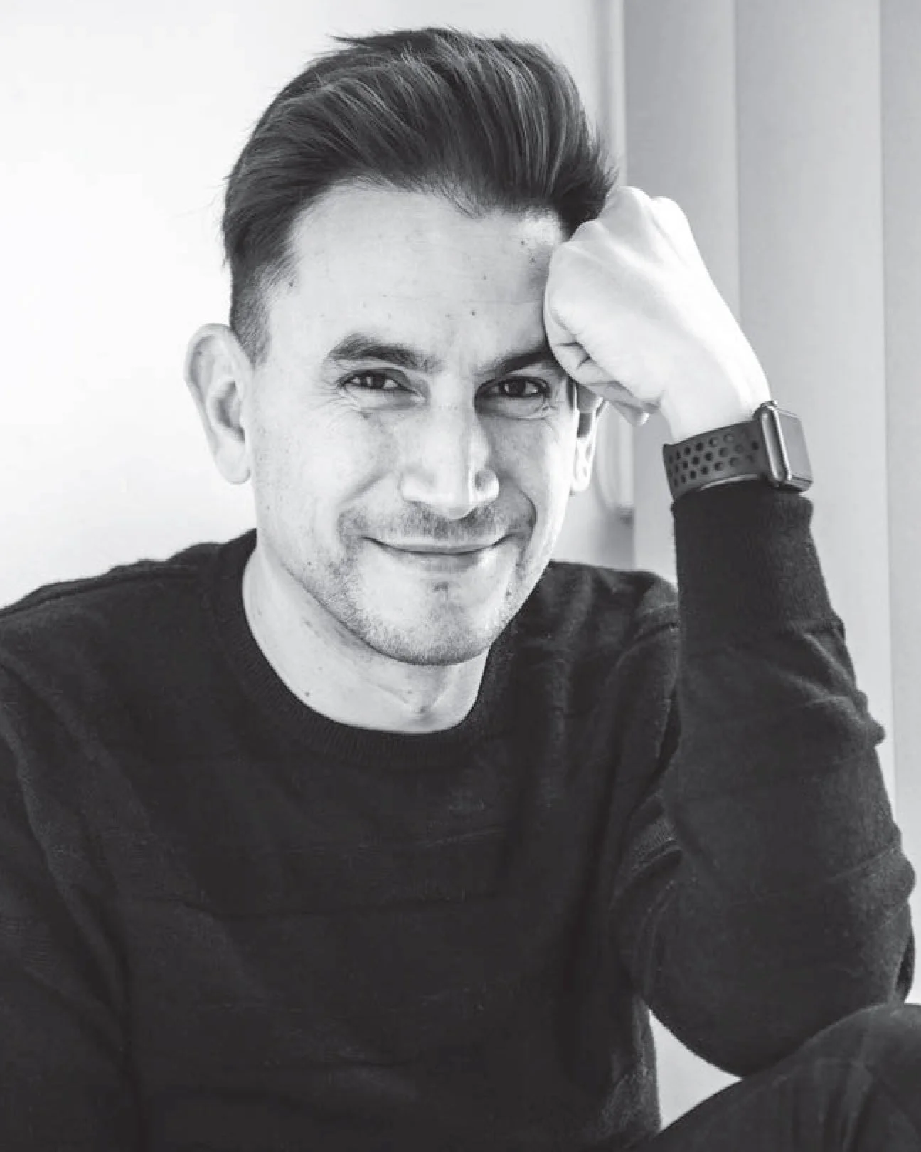 Black and white photo of a smiling man with dark hair, wearing a black sweater, and a smartwatch, sitting with his arm resting on his head.