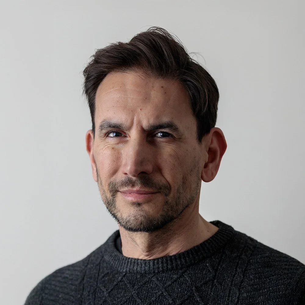 Black and white photo of a smiling man with dark hair, wearing a black sweater, and a smartwatch, sitting with his arm resting on his head.
