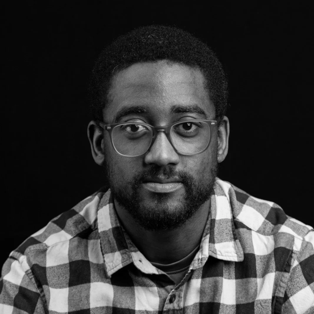 Black and white portrait of a man wearing glasses and a checkered shirt, looking directly at the camera against a dark background.