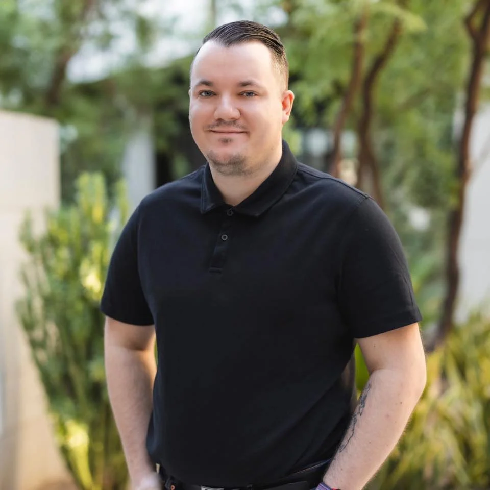 Person wearing a black polo shirt standing outdoors with greenery in the background.