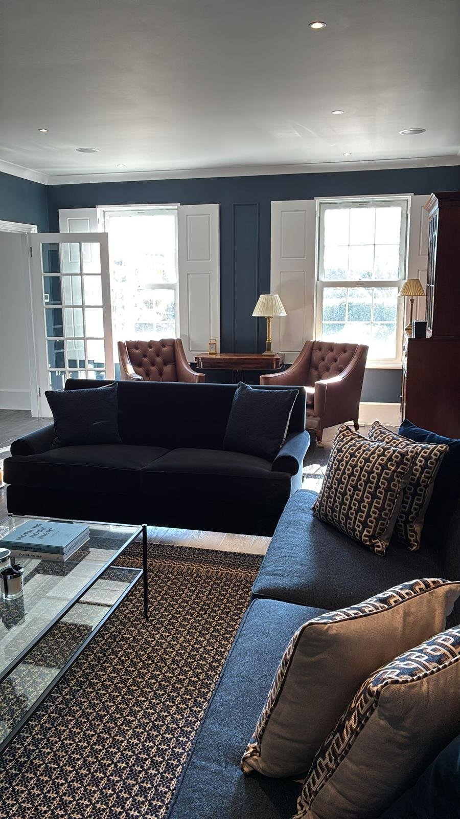 Living room with navy walls, two large windows with white shutters, a black sofa, a patterned rug, and a pair of brown armchairs near the windows with a wooden table and lamps between them.