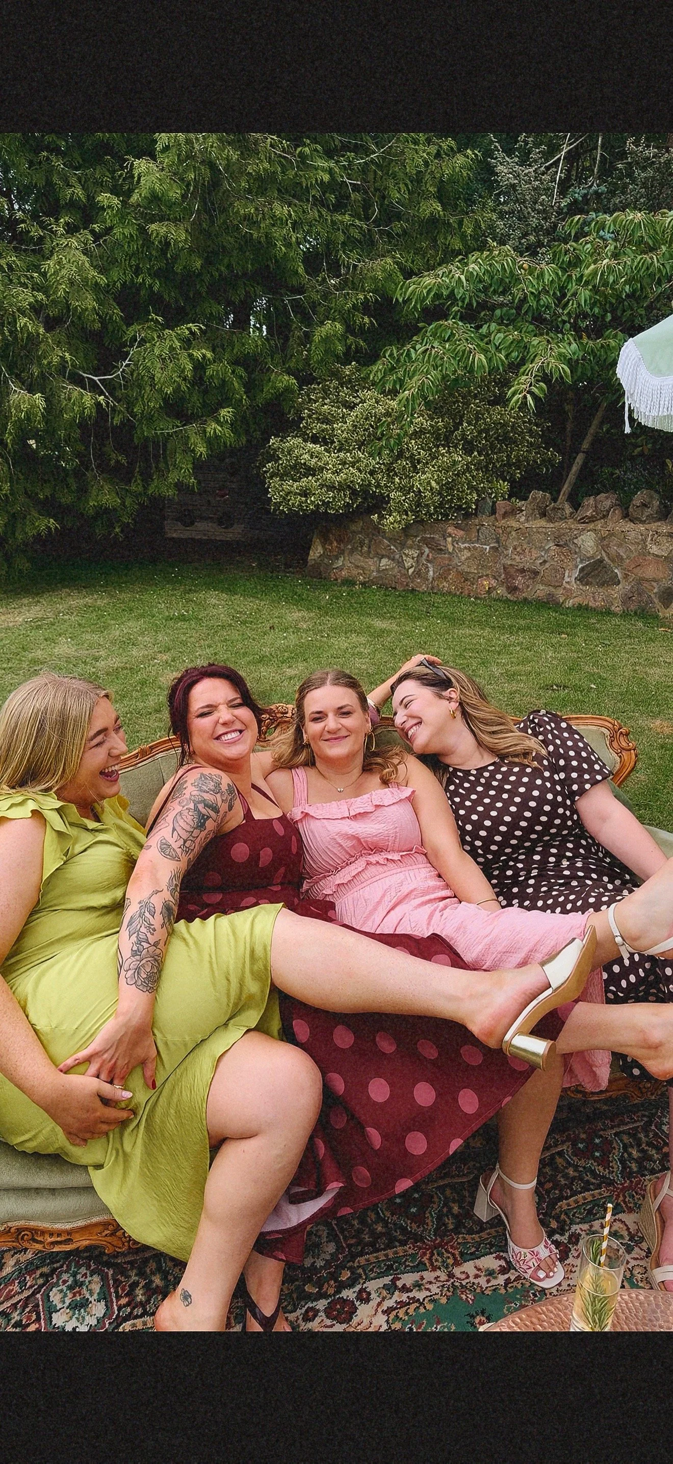 Four women sitting on a vintage couch outdoors, smiling and laughing, with greenery and trees in the background. Dudley wedding content creator.