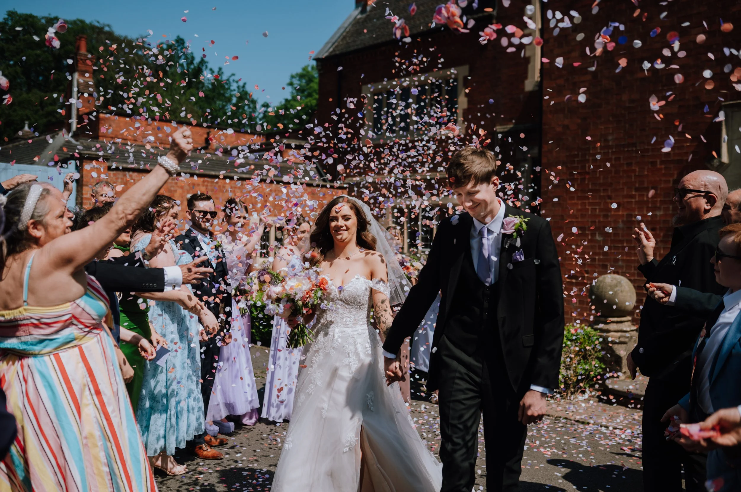 A bride and groom walking through a crowd of wedding guests throwing colorful confetti outside a brick building on a sunny day. Pendrell Hall Wedding.