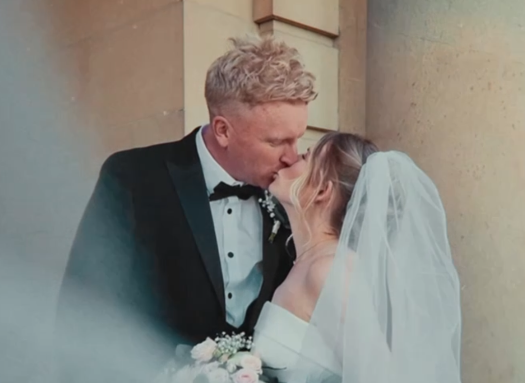 A newlywed couple shares a kiss at their wedding, with the groom in a tuxedo and the bride in a wedding dress and veil. Netley Hall Wedding.