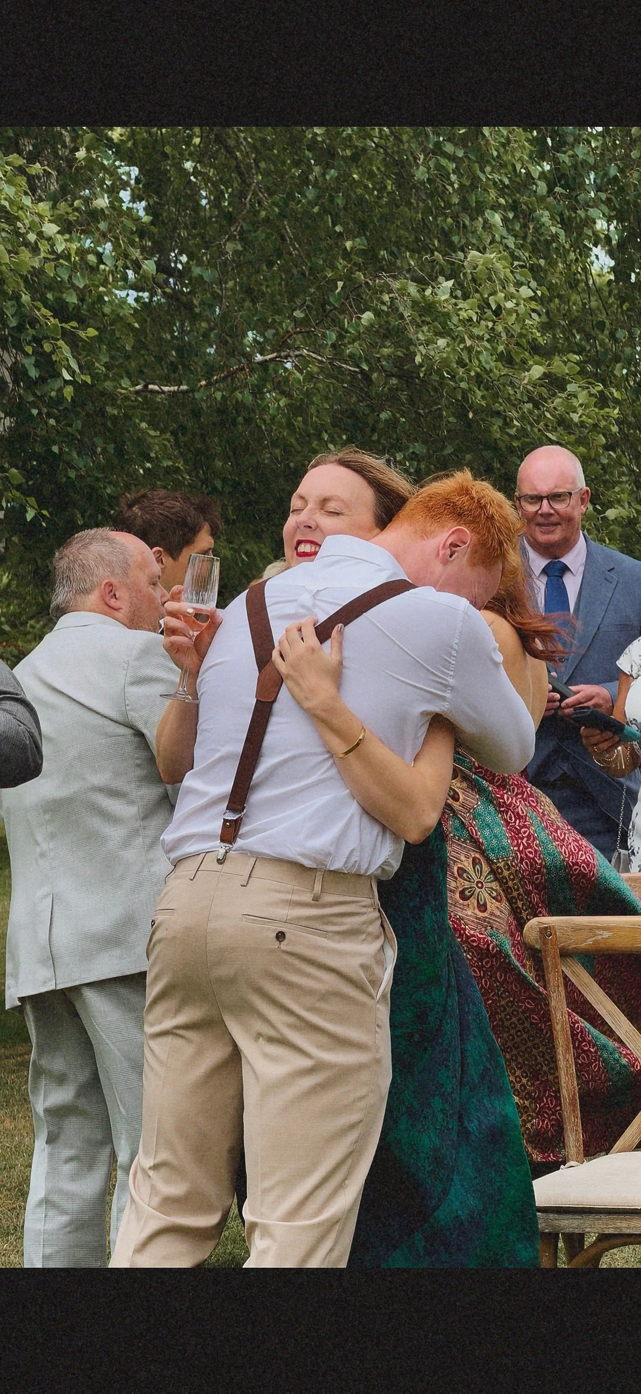 People hugging outdoors during a celebration, with several men and women smiling and holding drinks, in front of leafy trees. Dudley wedding content creator.