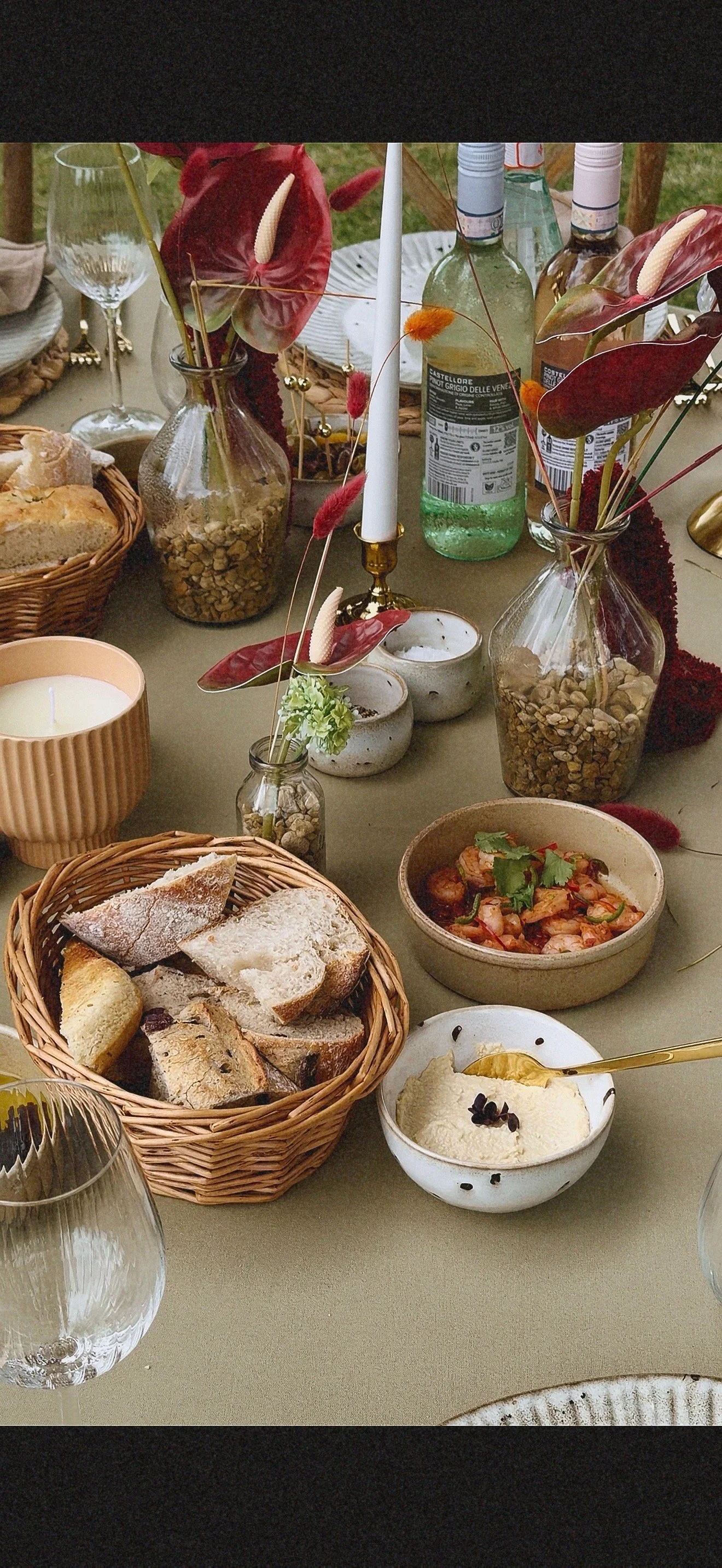 A table set with various food items including bread, a bowl of dip with herbs, a dish with shrimp, a small bowl of cheese or dip, a bread basket, candles, bottles, and decorative vases with flowers and leaves. Dudley wedding content creator.