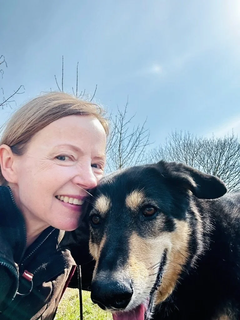 Eine Frau und ein Hund, die draußen im Freien posieren, mit blauen Himmel und Bäumen im Hintergrund.