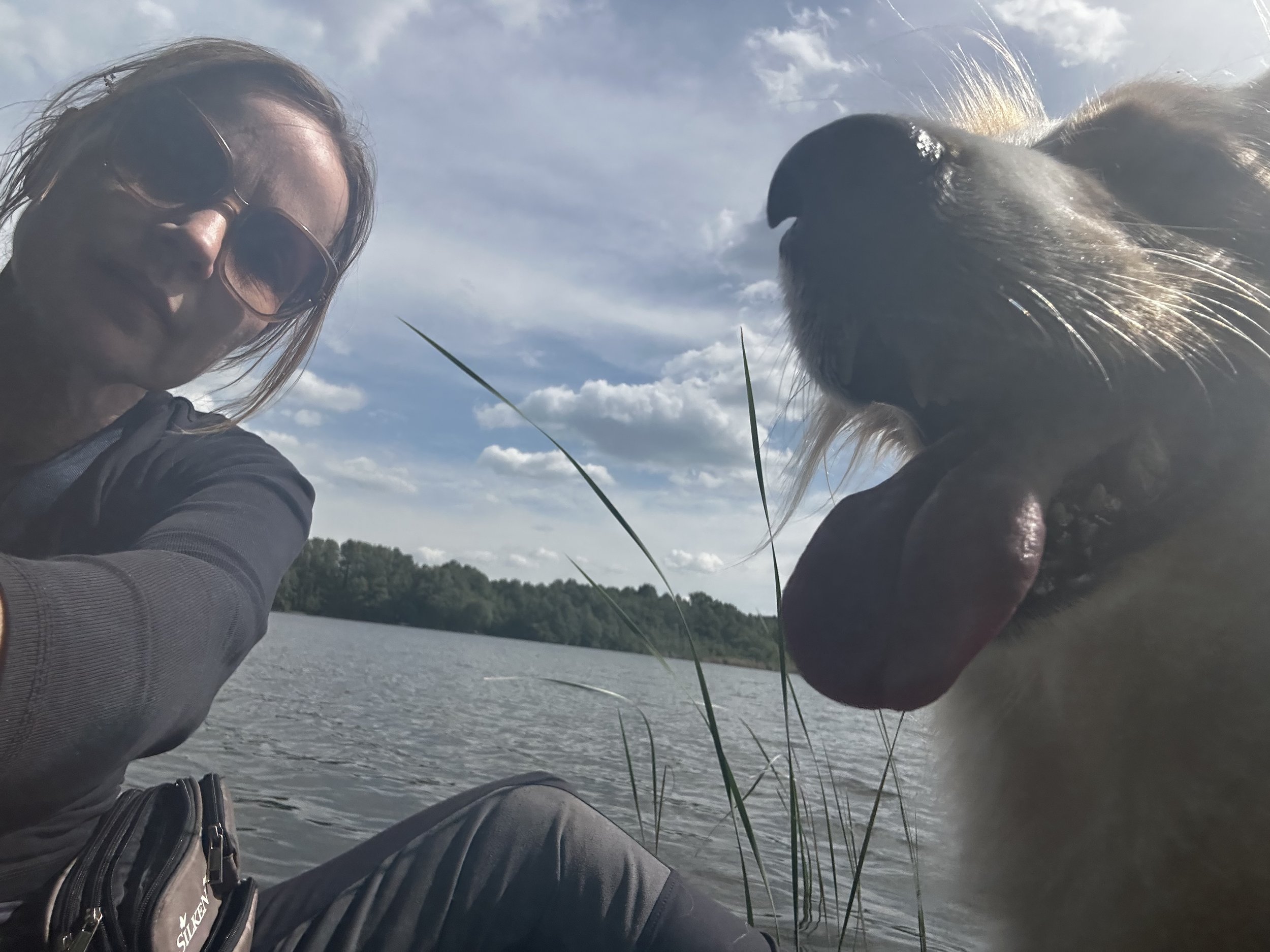 Frau mit Sonnenbrille und gestreifter Jacke, die einen See fotografiert, neben einem Hund mit herausgestreckter Zunge, bei sonnigem Himmel.