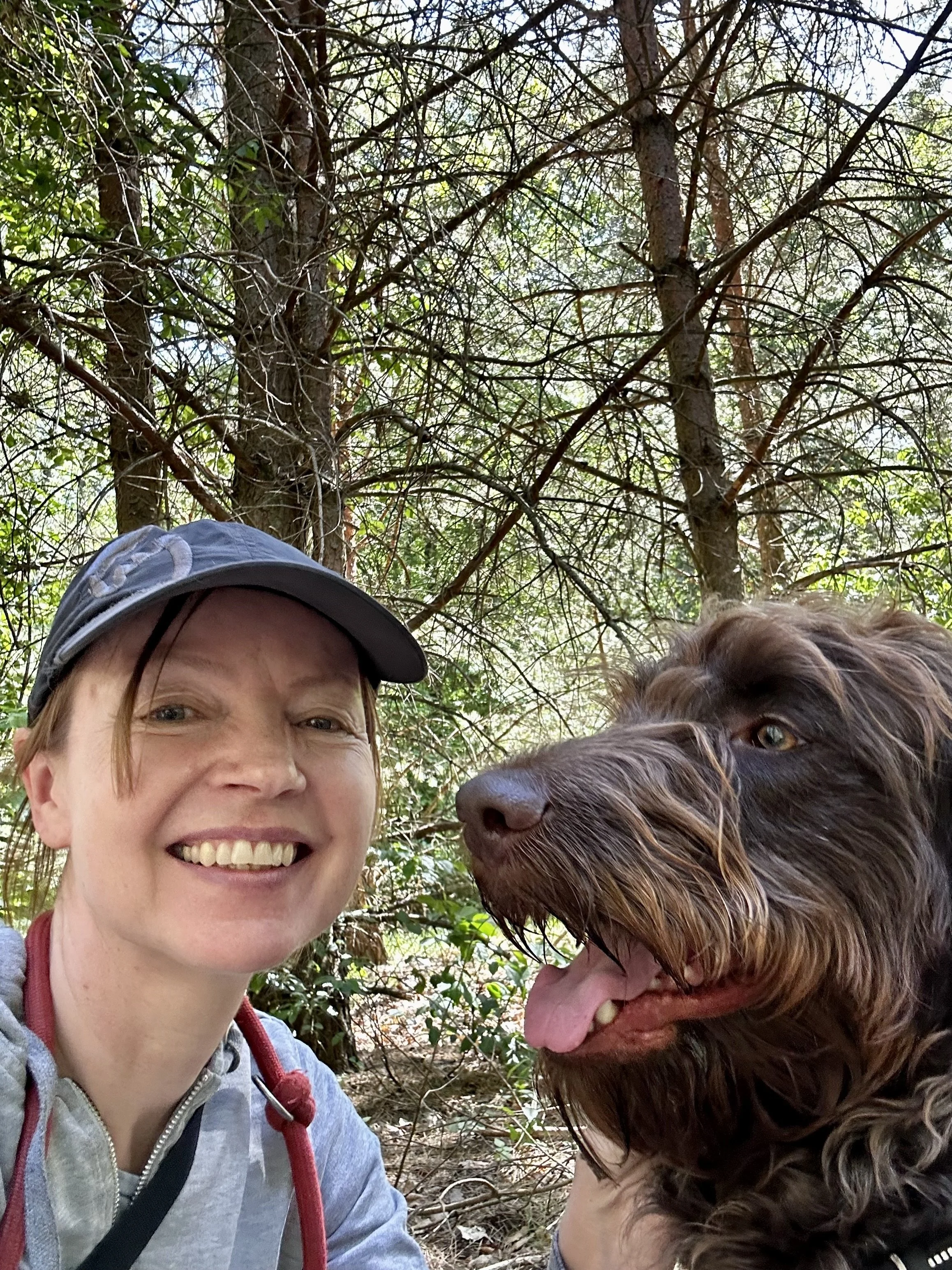 Frau mit braunem Hund in einem Wald, beide lächeln und schauen in die Kamera, umgeben von Bäumen und Flechten.