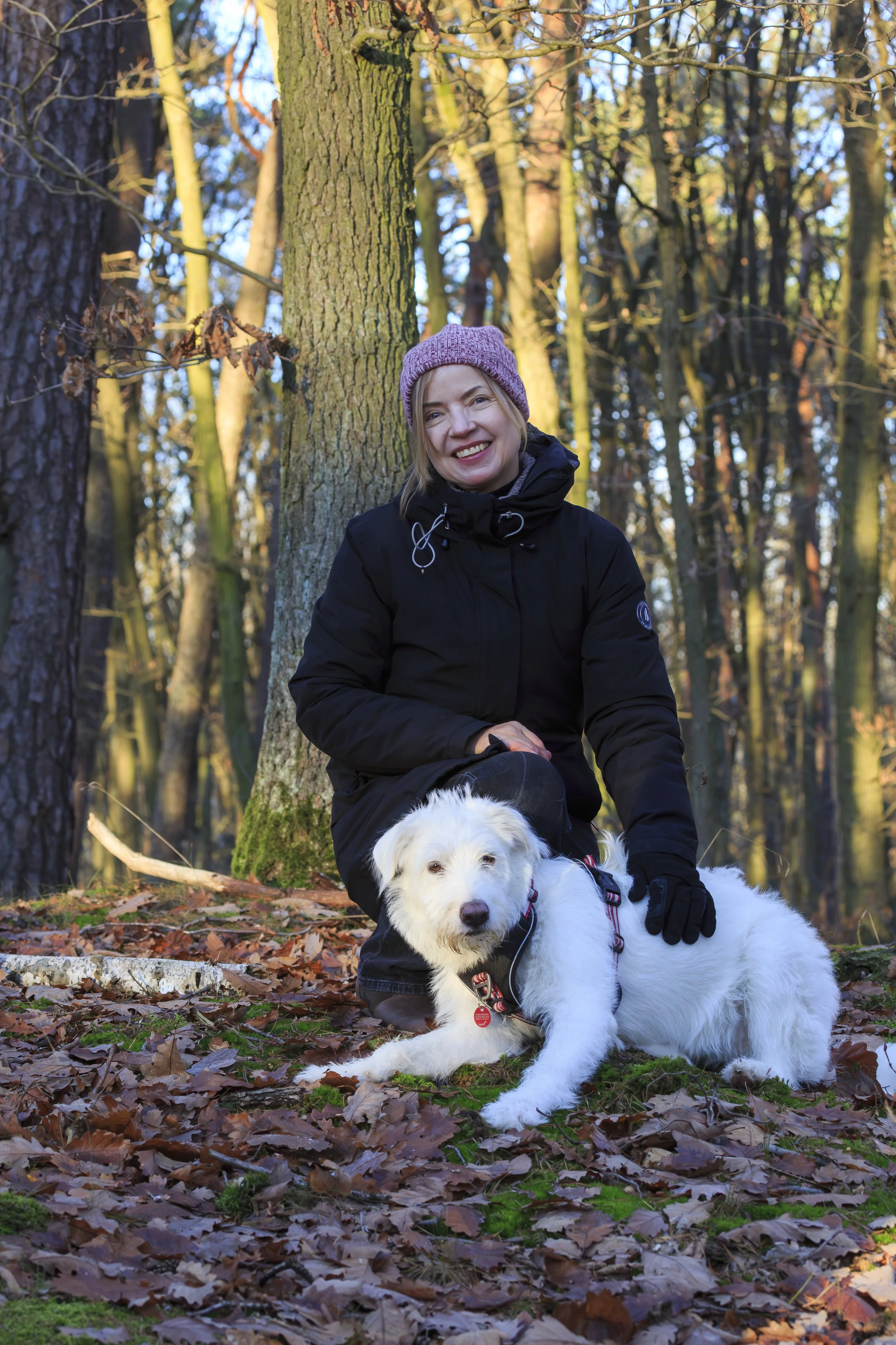Eine lachende Frau mit schwarzer Jacke und rosa Mütze im Wald, mit Weißem Hund auf Herbstlaub.