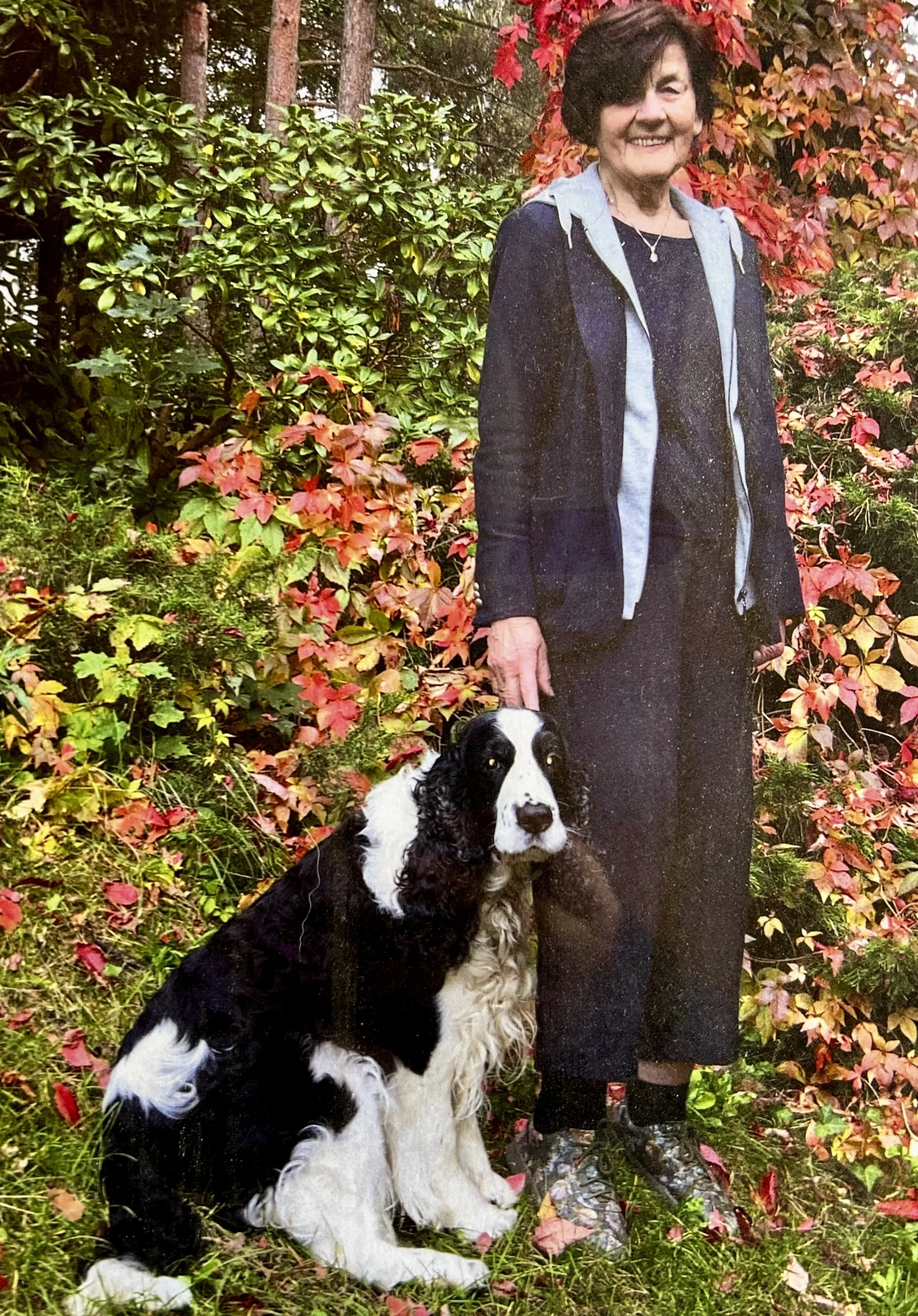 Eine ältere Frau steht im Garten neben einem schwarzen und weißen Hund, umgeben von bunten herbstlichen Blättern.