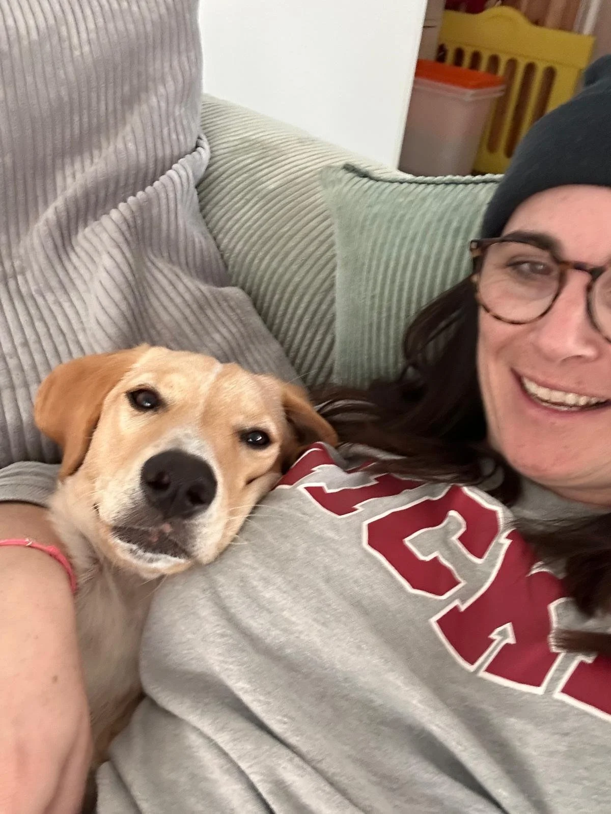 Eine Frau liegt auf einer Couch mit einem Hund, beide lächeln und schauen in die Kamera. Der Hund liegt nah am Gesicht der Frau, auf ihrem Oberteil.