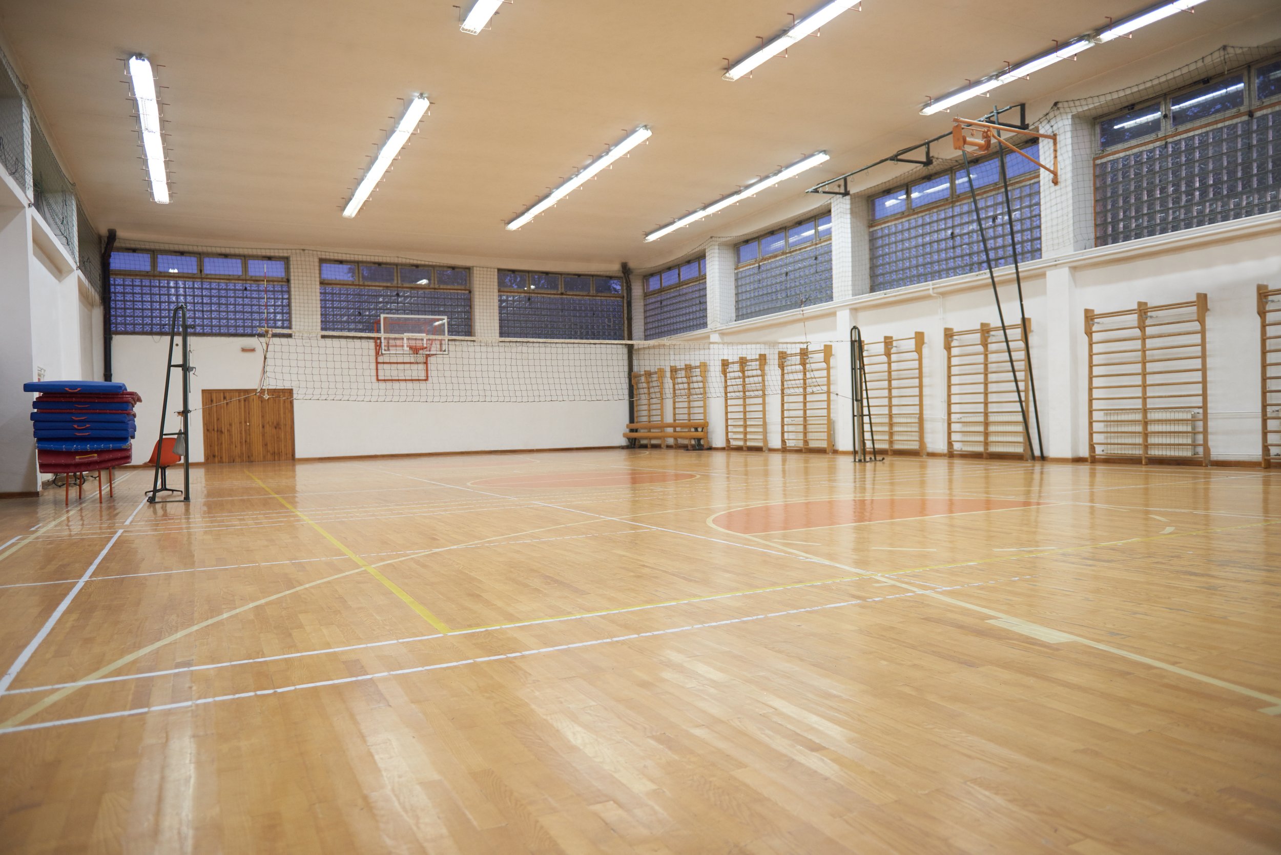 Empty indoor gymnasium with wooden flooring, volleyball net, basketball hoop, foldable mats stacked on chairs, and wall-mounted exercise bars.