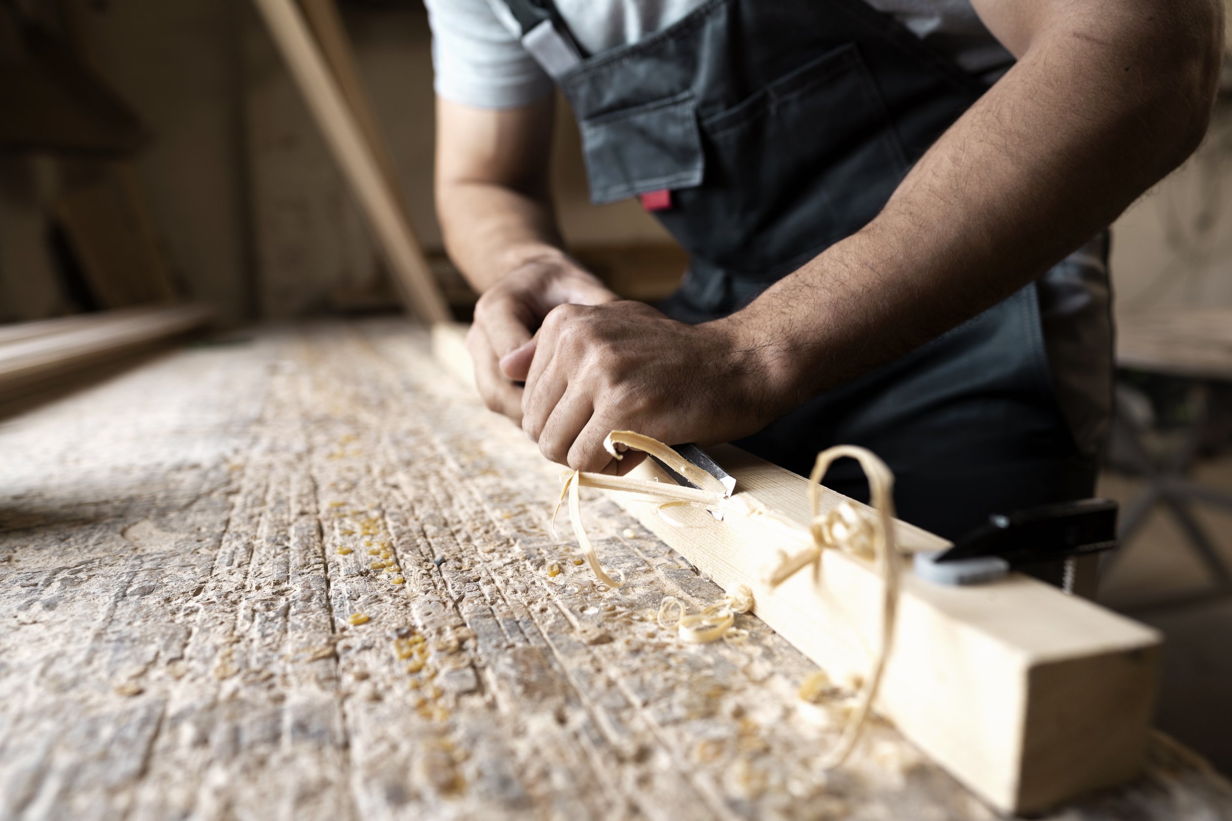 A person using a hand saw to cut a piece of wood in a woodworking shop, with wood shavings on the work surface.