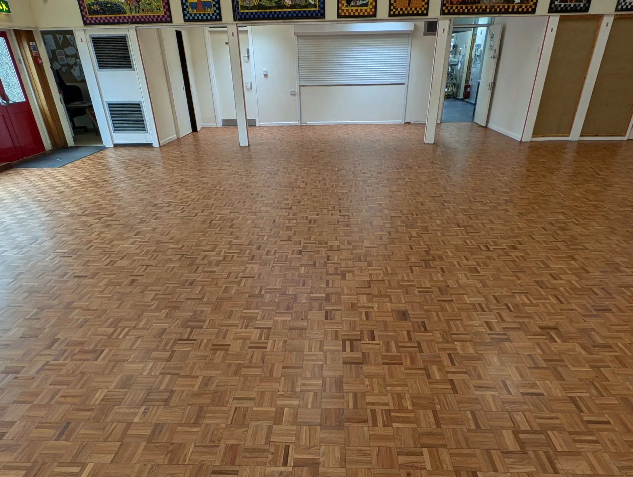 Empty room with wood parquet flooring, beige walls, and several doors and windows, some framed with wood. There are some bulletin boards and a red door on the left side.