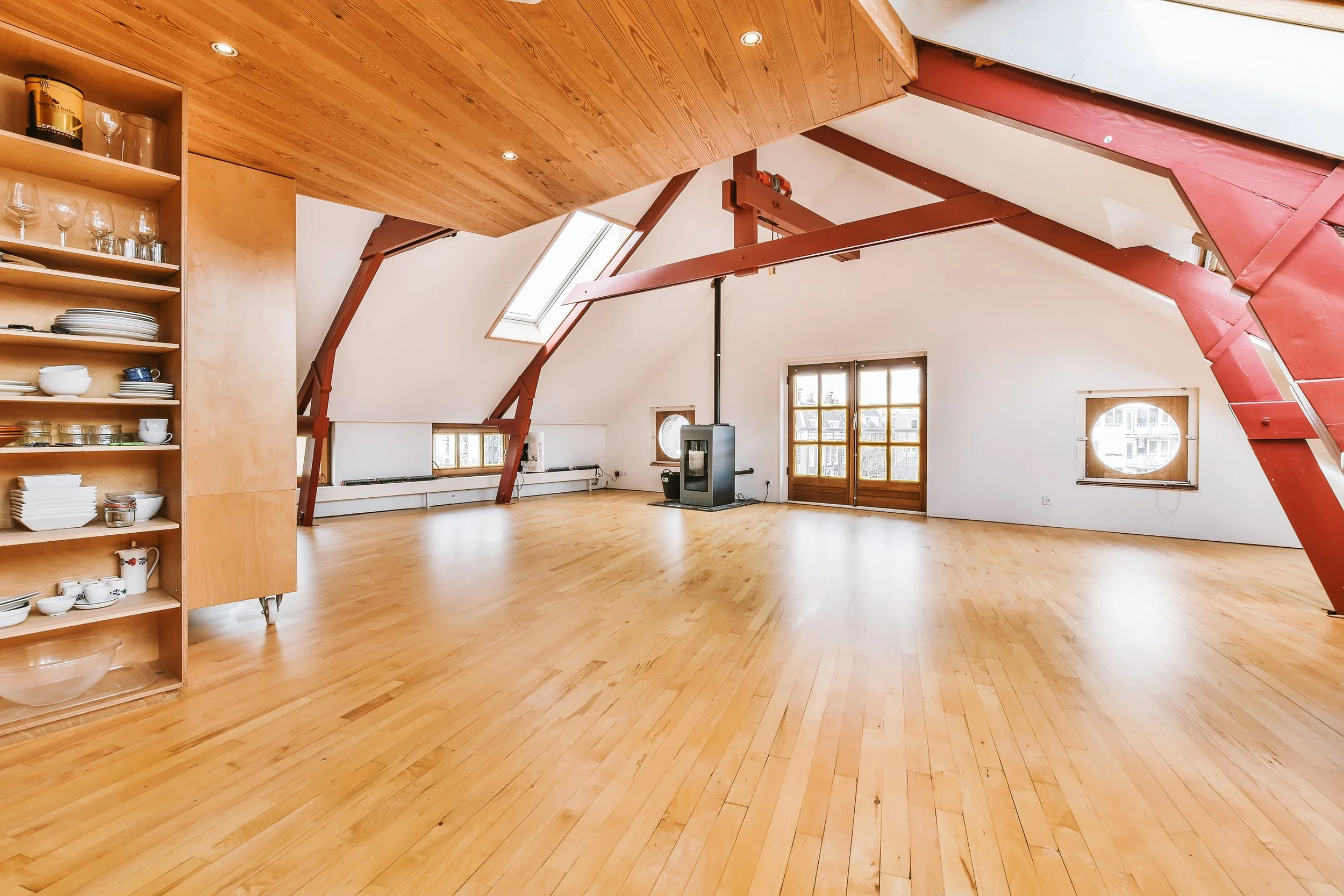 Spacious attic room with wooden floor, sloped ceiling, red beams, large windows, and a wood-burning stove.