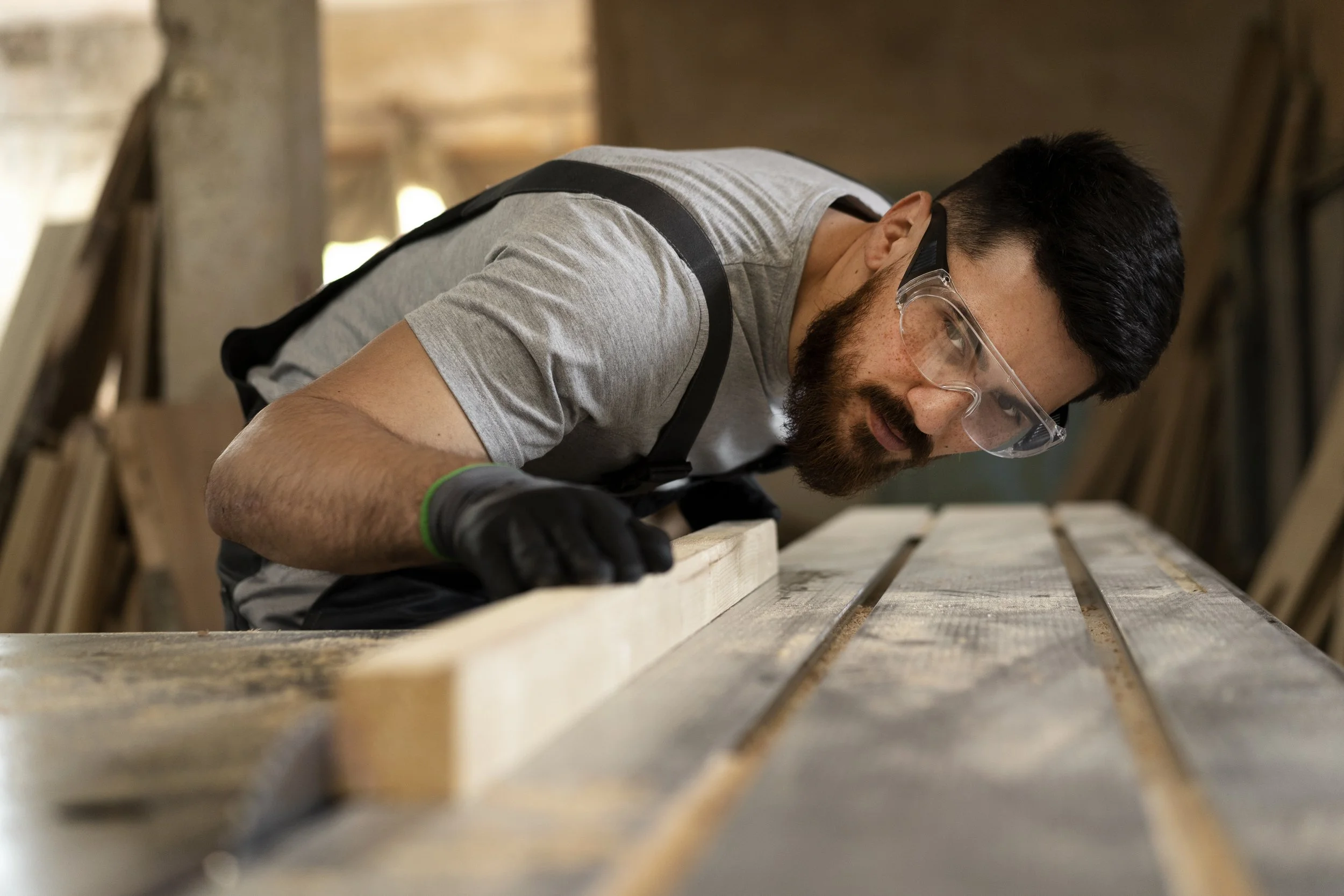 A man with safety glasses and gloves is sanding a wooden plank in a woodworking shop.