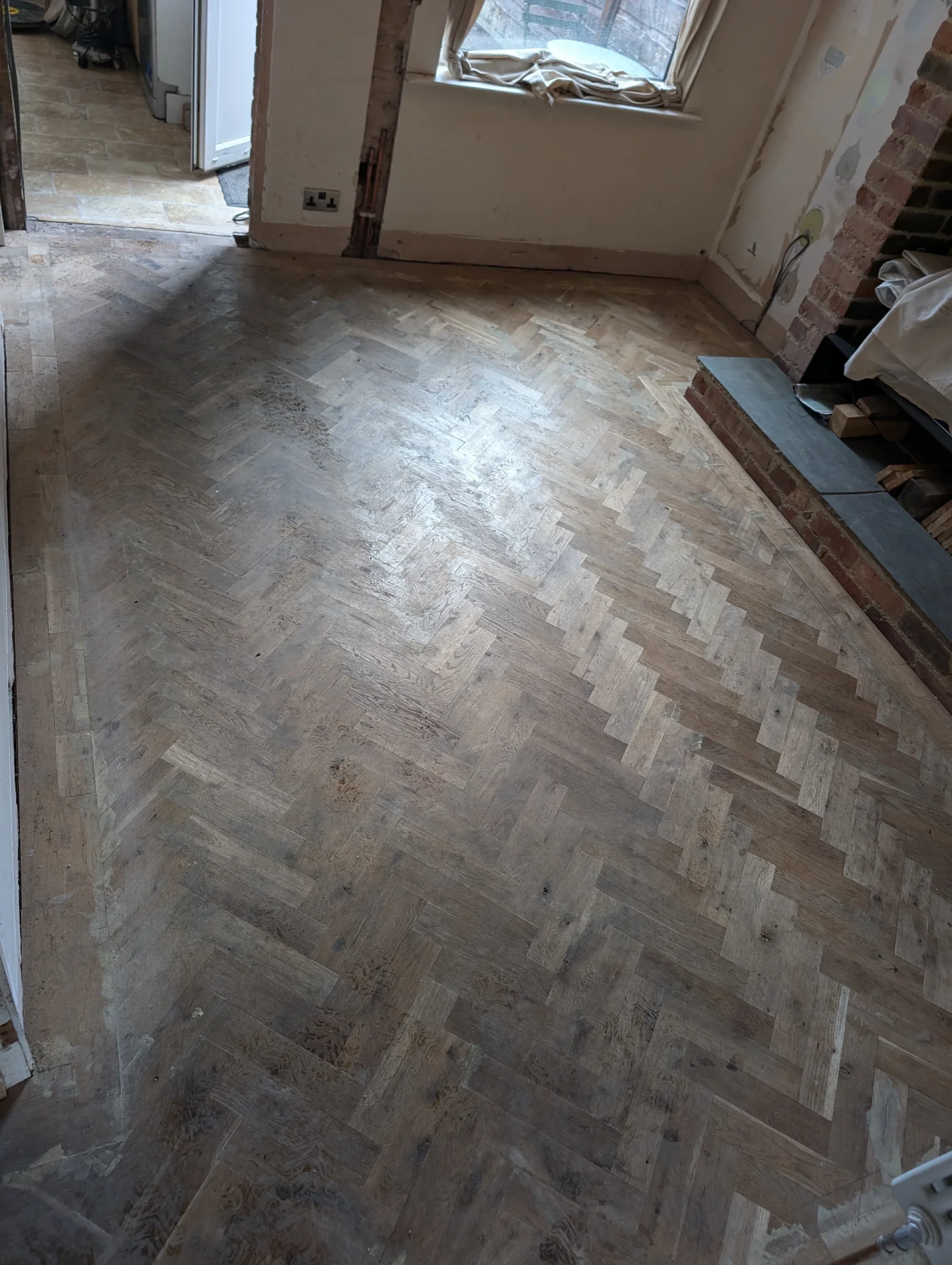 Wooden herringbone floor being installed in a room under renovation, with a fireplace and a window in the background.