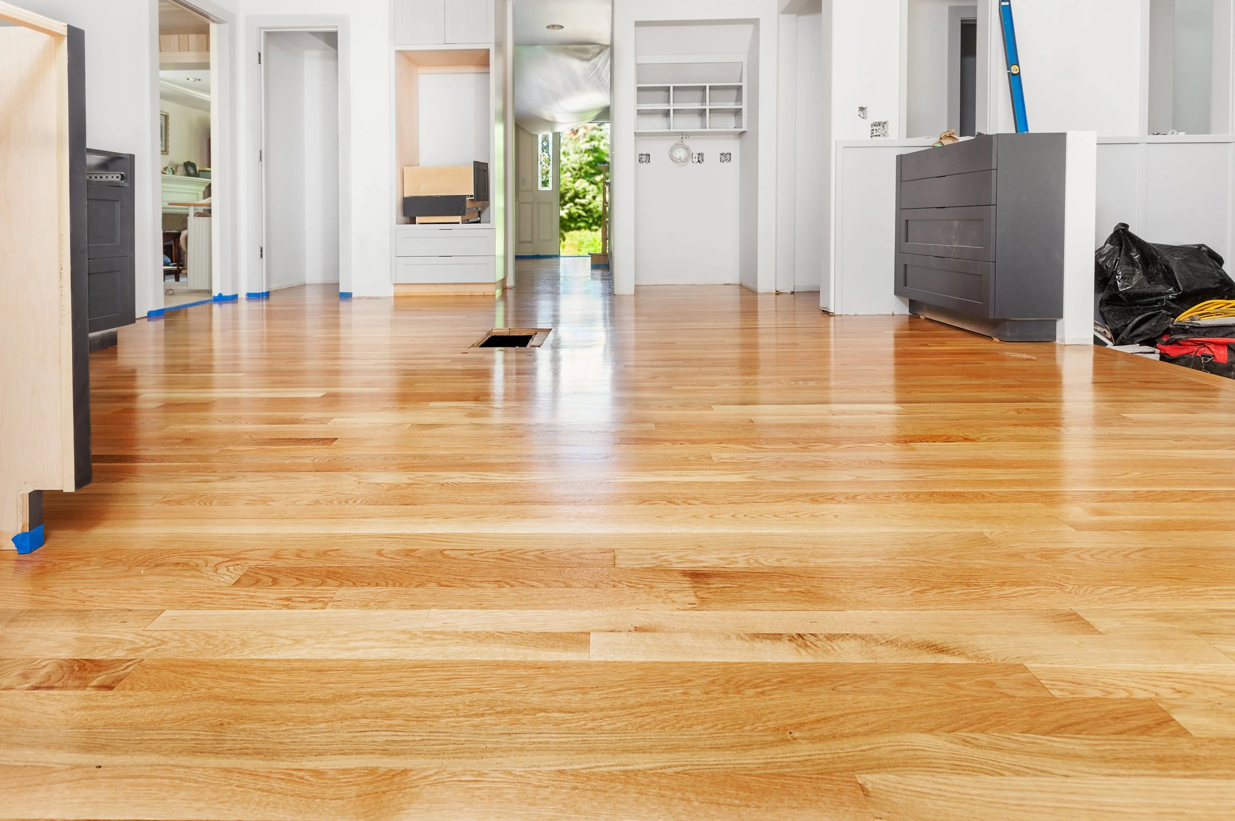 Empty room with freshly installed hardwood floors, white walls, and some construction tools and materials, with an open front door revealing greenery outside.
