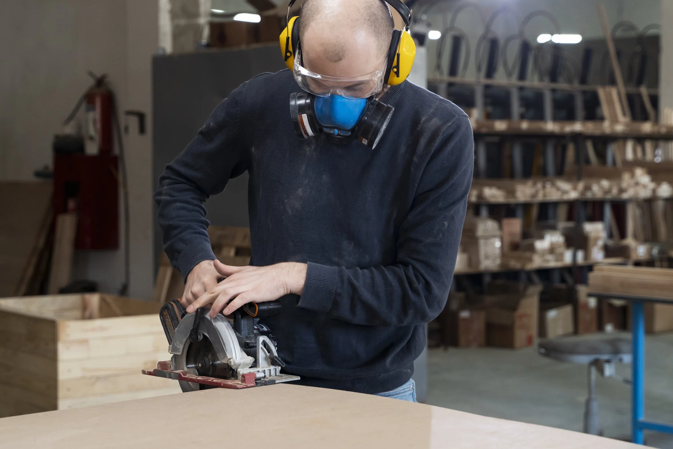 A person wearing a dust mask, yellow ear protection, and safety goggles is using a circular saw to cut a piece of wood in a woodworking shop.
