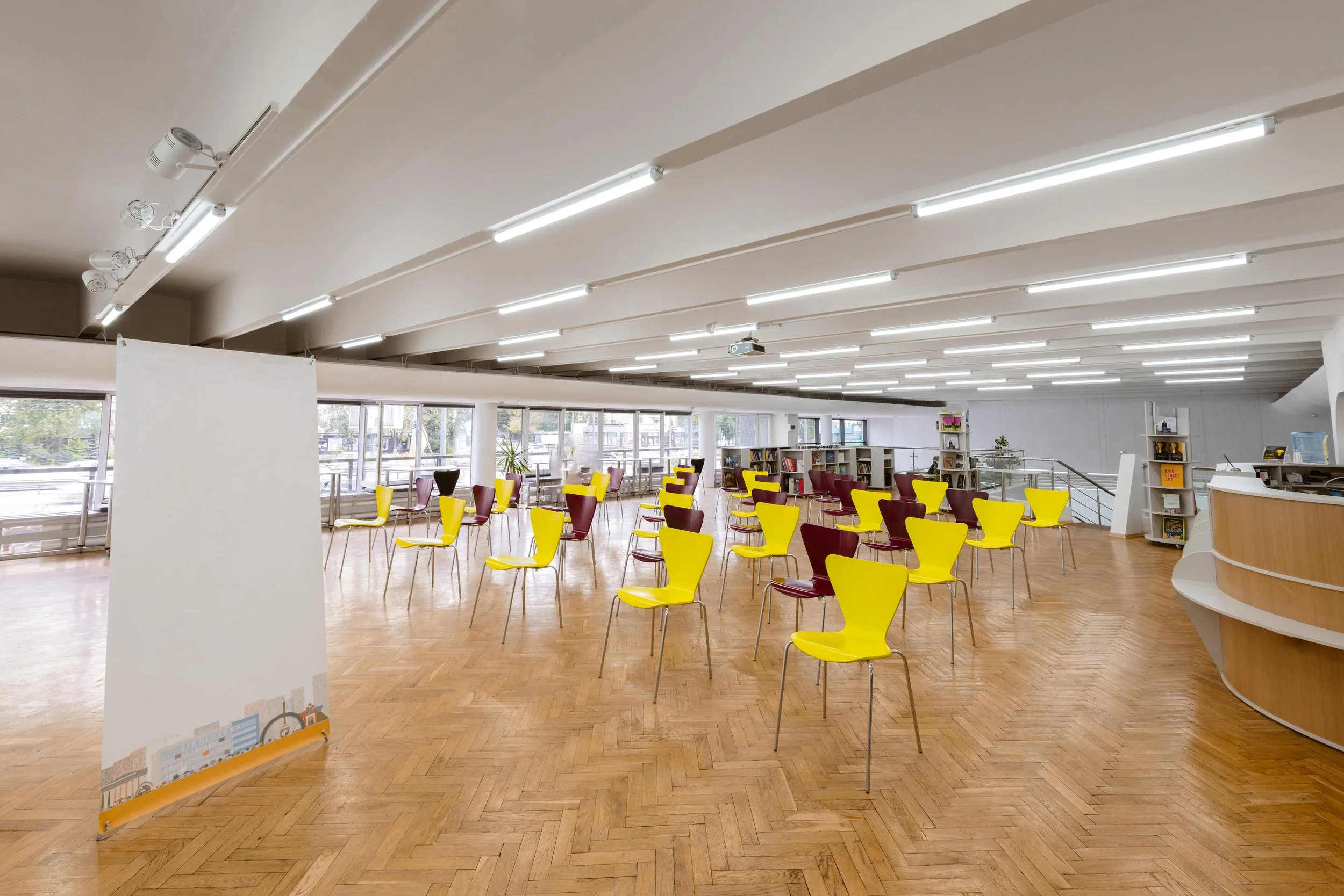 An open, well-lit room with yellow and burgundy chairs spaced out, likely set up for an event or seating arrangement. Large windows, bookshelves, and a curved reception desk are visible in the background.