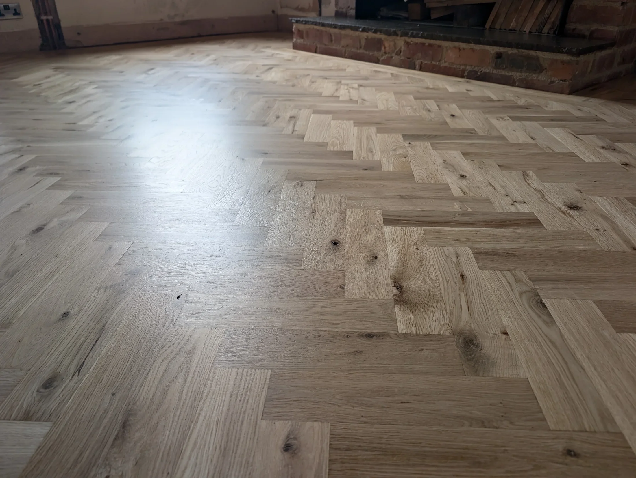 Close-up of a newly installed wooden parquet floor with a herringbone pattern near a brick fireplace.