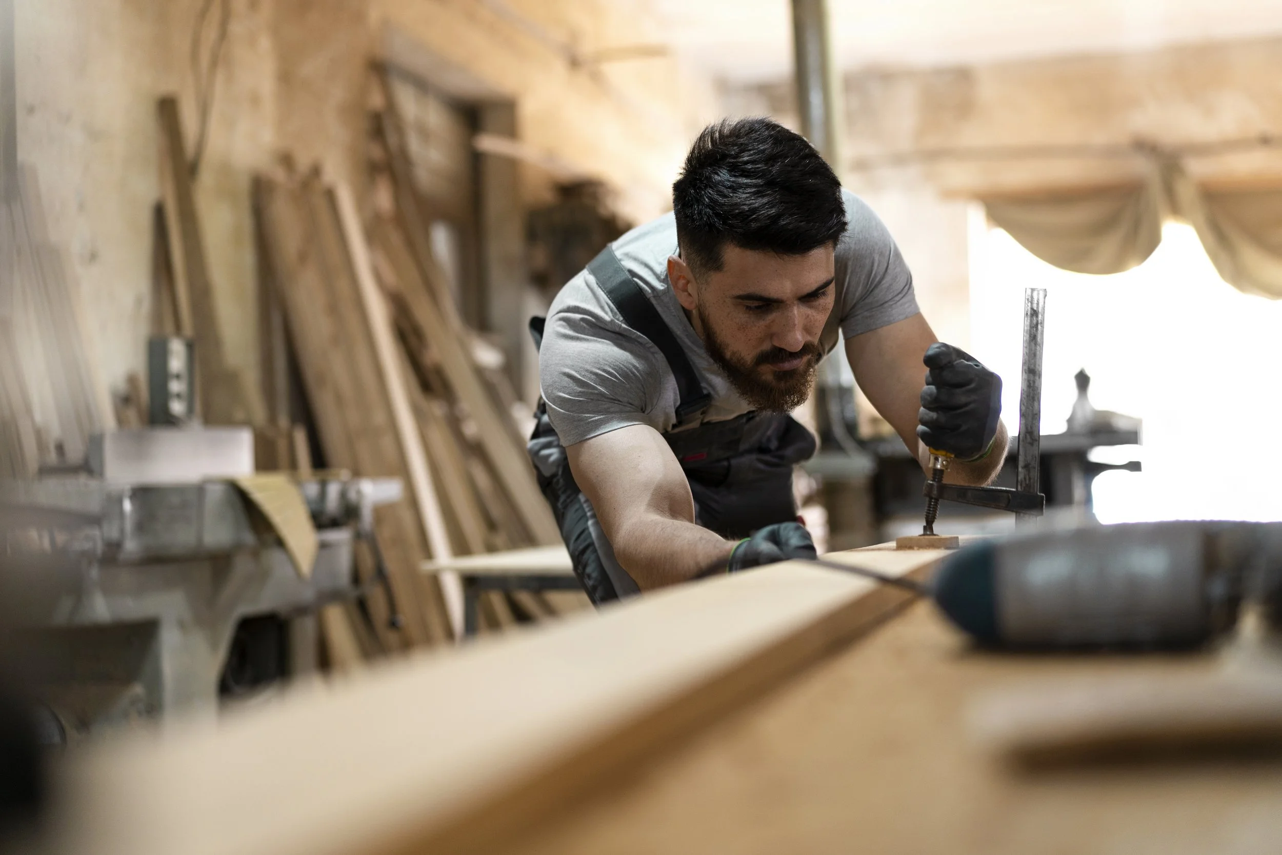 A man working in a woodworking shop, using a clamp on a piece of wood. The background shows various pieces of wood and tools.