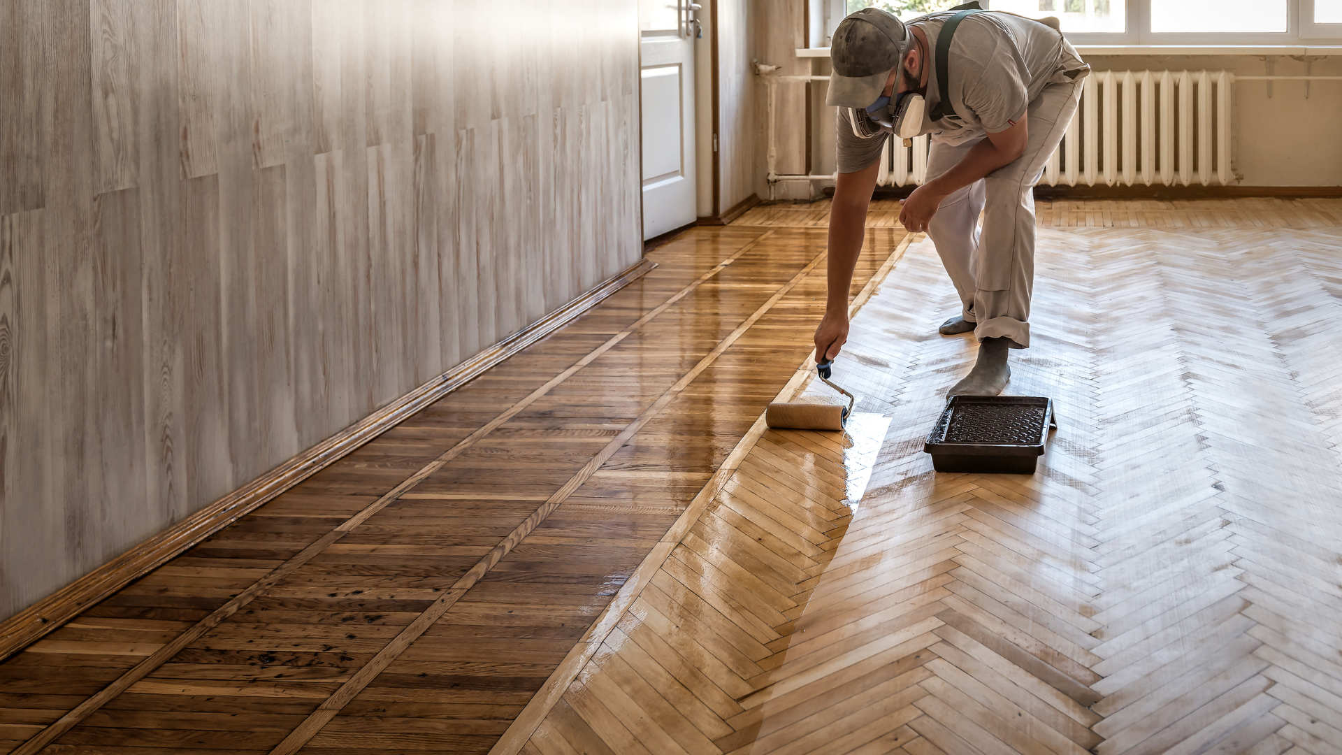 A person applying a finish to a wooden floor using a paint roller, with a tray of finish nearby, in a room with wooden paneling and a radiator.