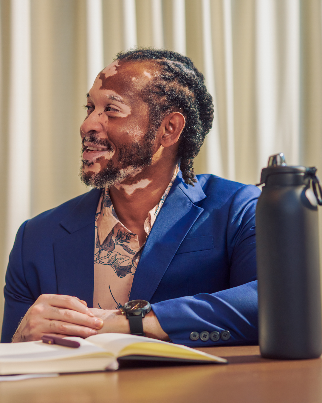 A Black man with vitiligo sits at a desk, smiling to someone off-camera. He wears a navy blazer with a peach dress shirt.