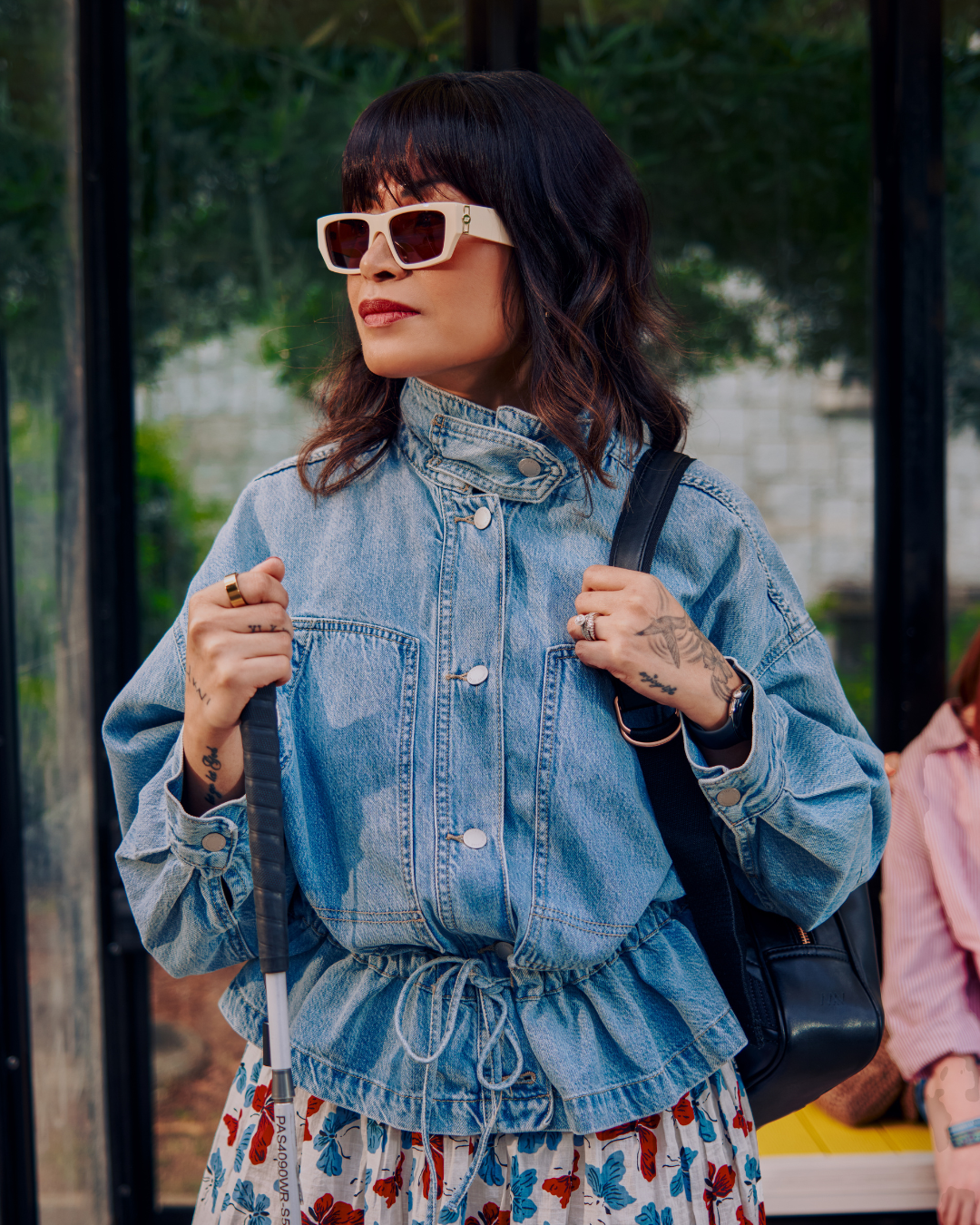 A Black woman with a visual impairments stands waiting at a bus shelter holding her cane. She wears a jean jacket, sunglasses, and a floral skirt.