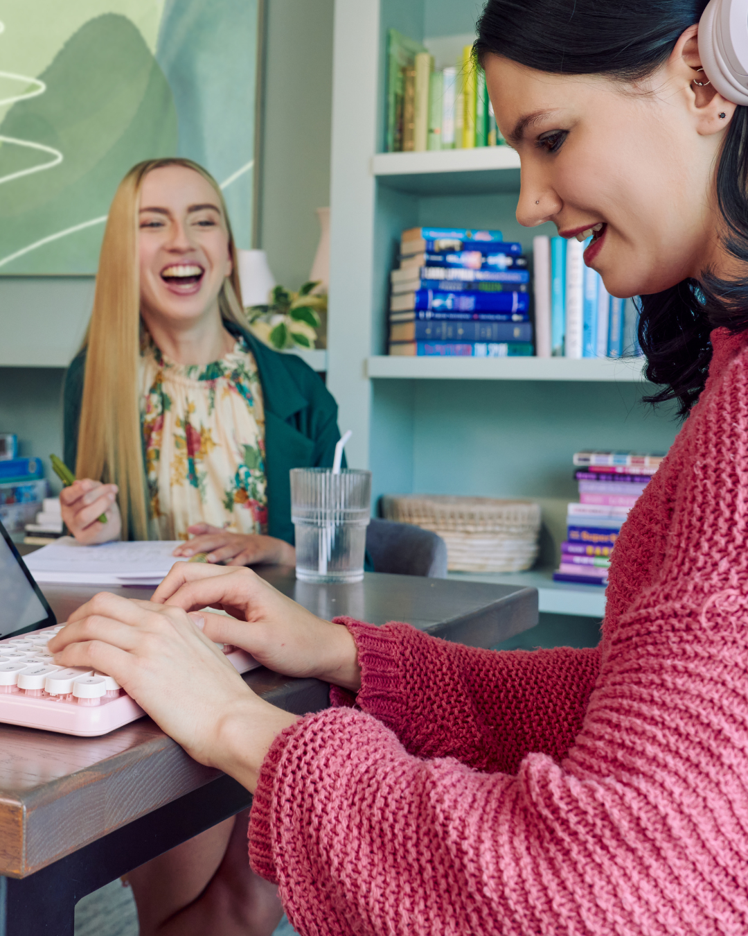 Two white women sit chatting at a shared desk, co-working. The woman in the foreground is using an adaptive keyboard.