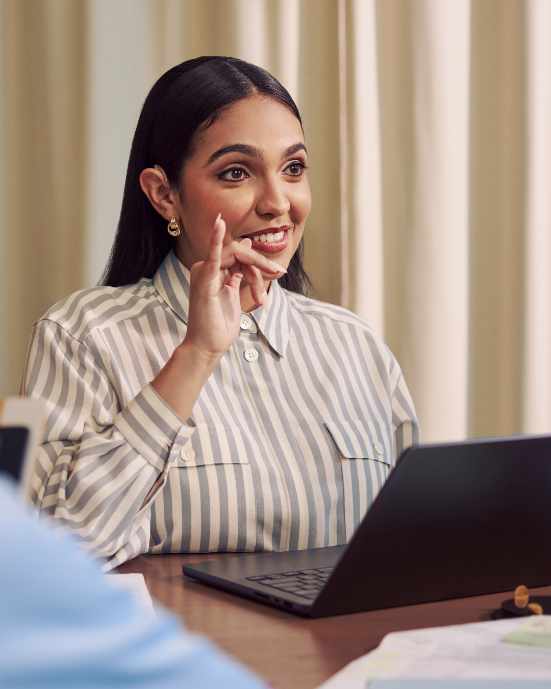 A Black woman signs ASL to her interpreter seated off camera. She wears a white and grey striped shirt and sits at a desk with a laptop