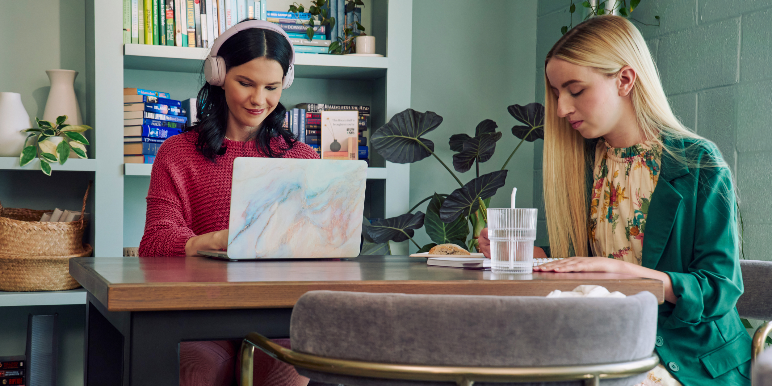 A young pale white blind woman and a young white woman with partially paralyzed arms and hands are coworking at a table in a modern coworking office space.