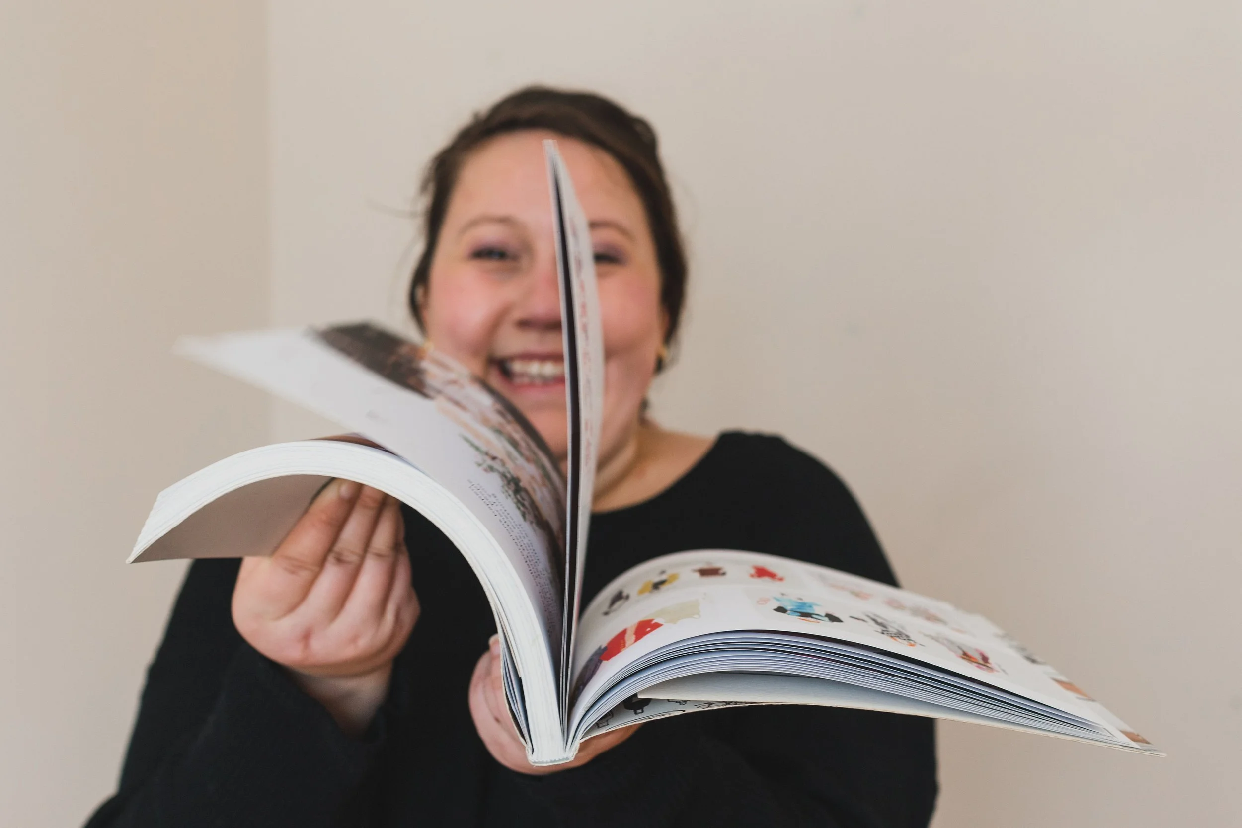 A young white woman in her thirties smiles at the camera. Her face is partially covered by a book she holds out in front of her while she flips through the pages