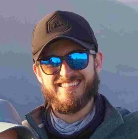 Smiling Hike Tribe's client with a beard wearing a cap and sunglasses, smiling outdoors.