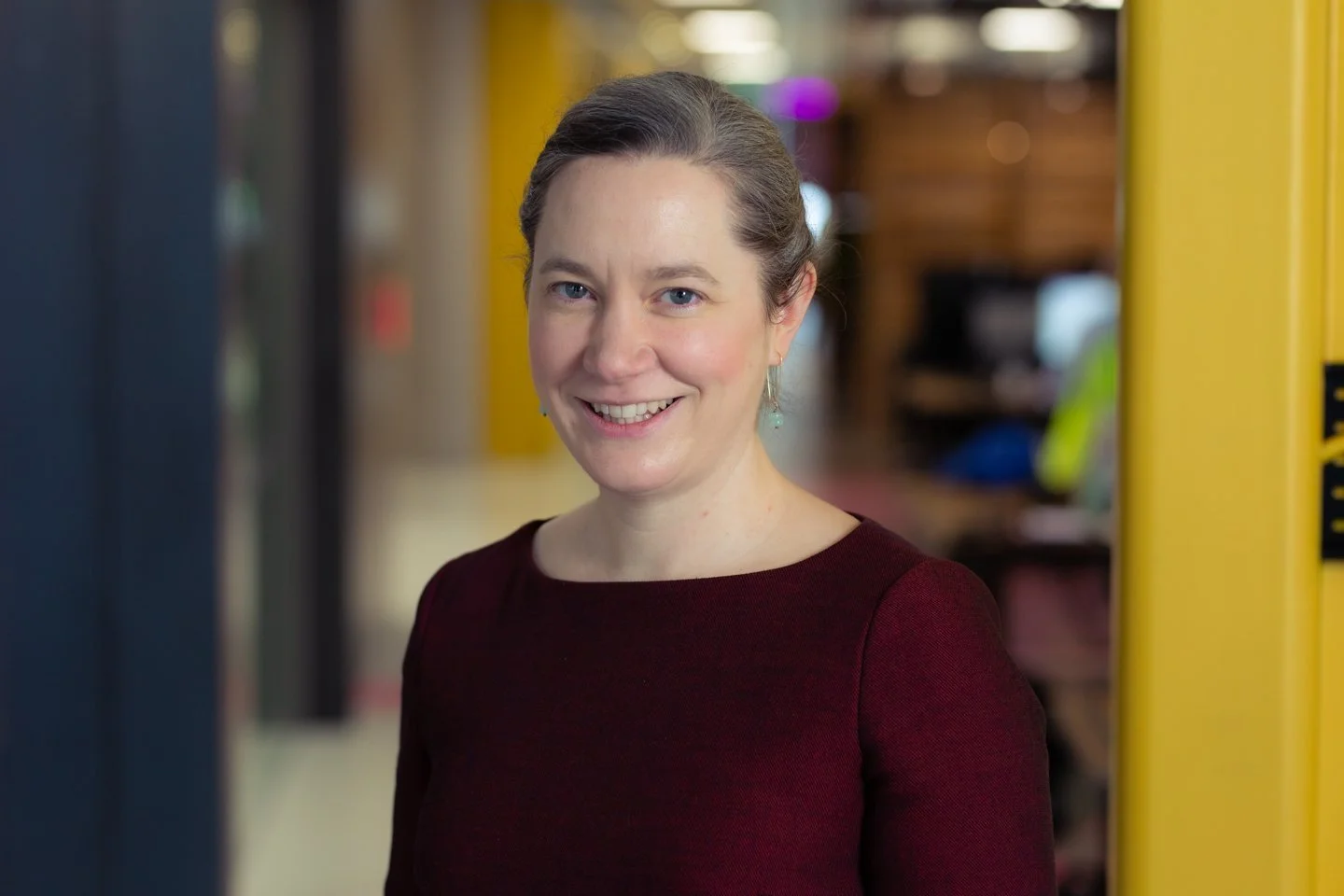 A woman with brown hair pulled back, smiling, wearing a dark red or burgundy long sleeve top, standing in an indoor setting with a yellow column and blurred background.